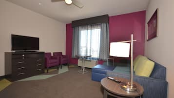 A hotel room with a flat-screen TV, a sofa, a small table with a lamp, a ceiling fan, and a window with patterned curtains.