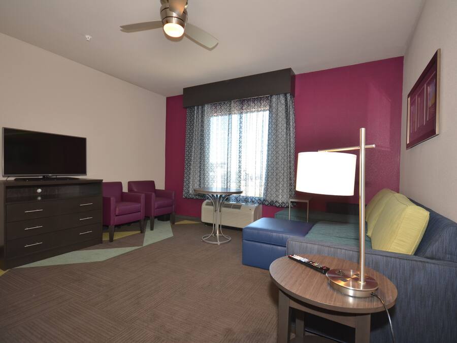 A hotel room with a flat-screen TV, a sofa, a small table with a lamp, a ceiling fan, and a window with patterned curtains.