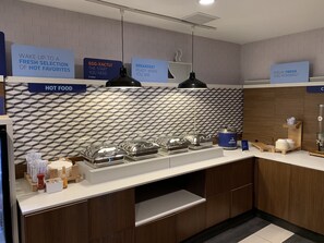 A modern breakfast bar with a geometric backsplash, labeled signs for hot food and breakfast, and a variety of breakfast items. - Holiday Inn Express & Suites Oklahoma City Mid - Arpt Area by IHG (Oklahoma City)