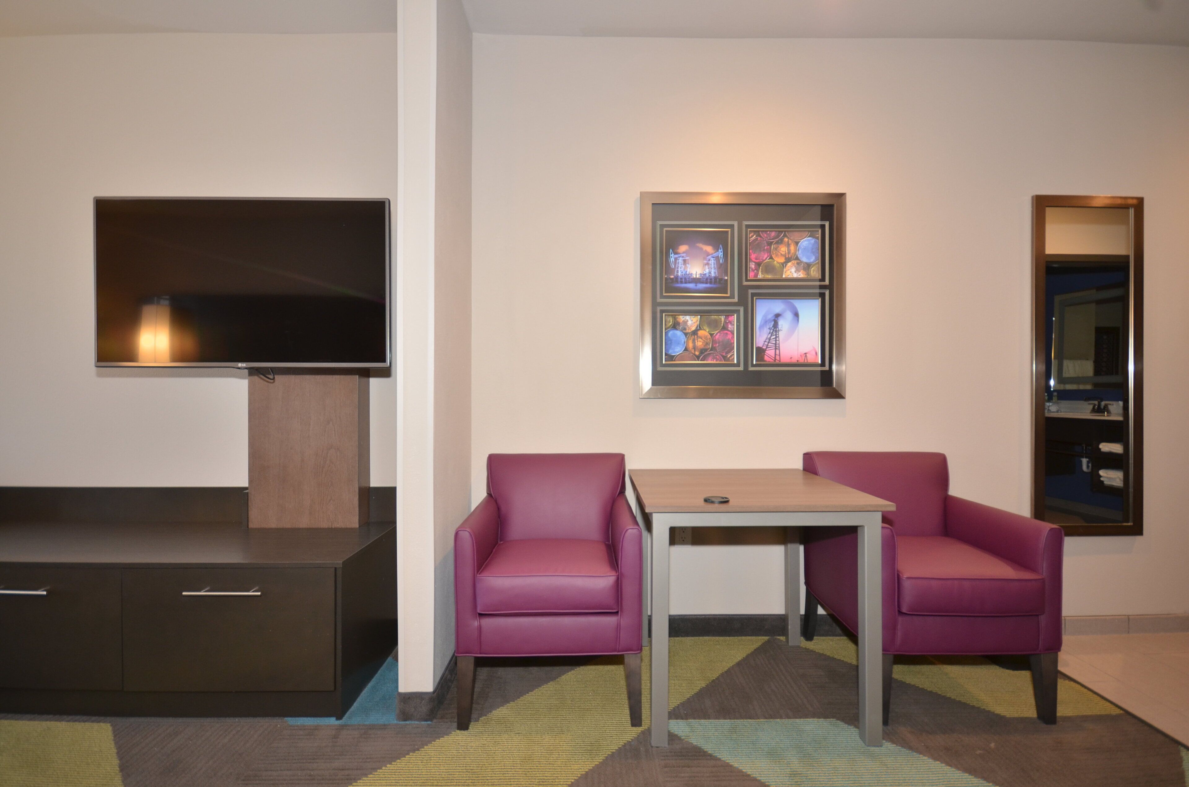 A hotel room with a flat-screen TV, a small table, two chairs, and a wall-mounted artwork.