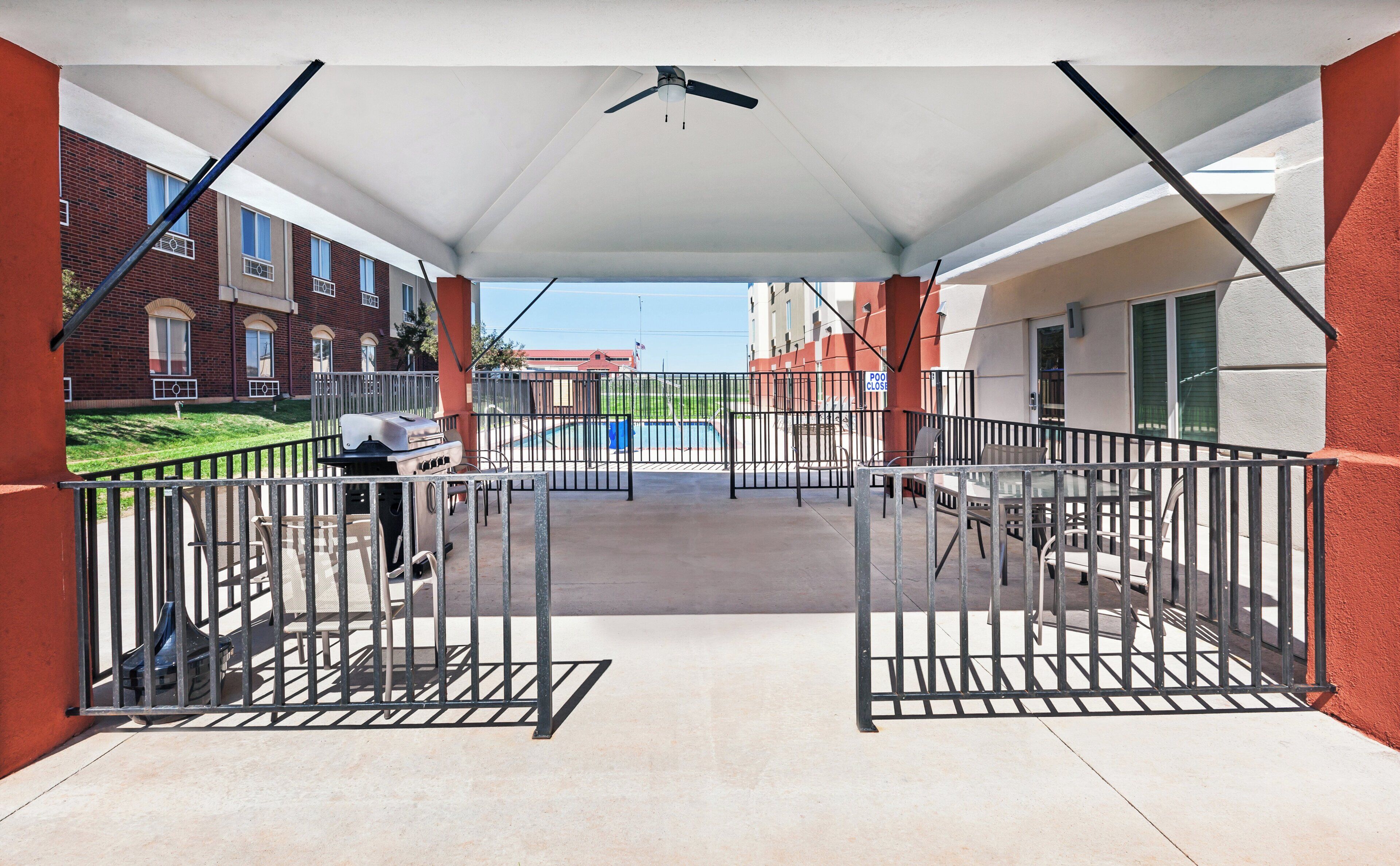 A covered outdoor area with a grill, seating, and a view of a swimming pool and buildings.