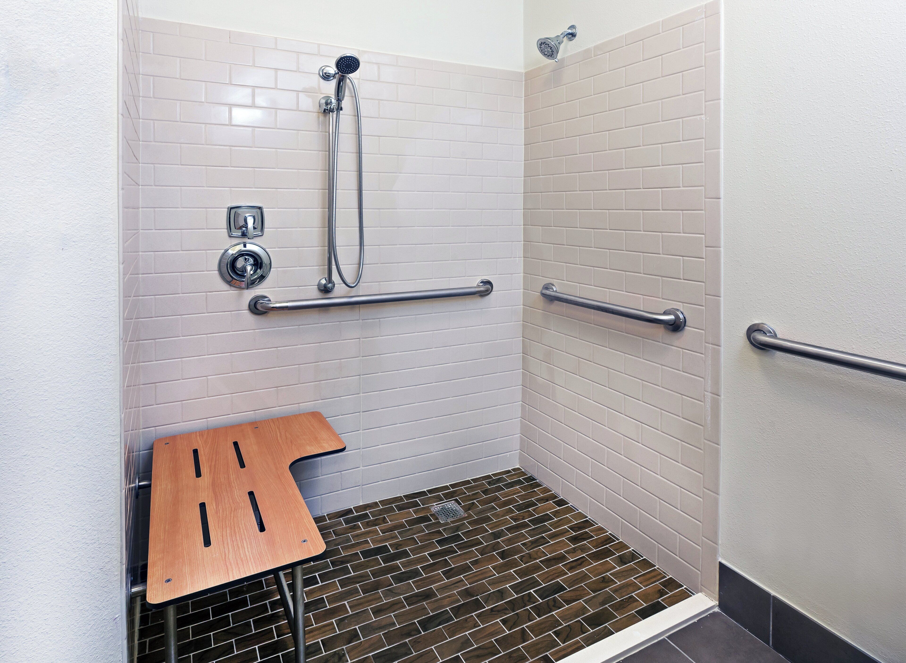 A shower area with white tiled walls, a handheld showerhead, a handheld shower attachment, and a grab bar.
