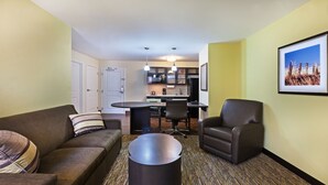 A compact living space with a sofa, armchair, and a coffee table. A kitchen area is visible in the background with a dining table and chairs. - Candlewood Suites San Angelo TX by IHG (San Angelo)