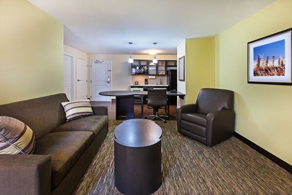 A compact living space with a sofa, armchair, and a coffee table. A kitchen area is visible in the background with a dining table and chairs.