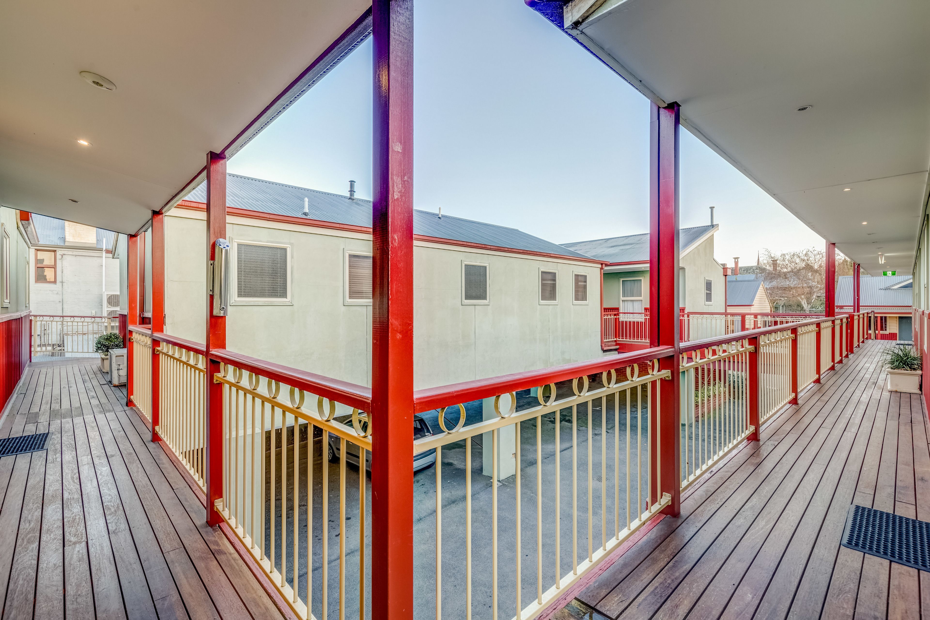 A covered outdoor area with a wooden deck, red railings, and a view of adjacent buildings.