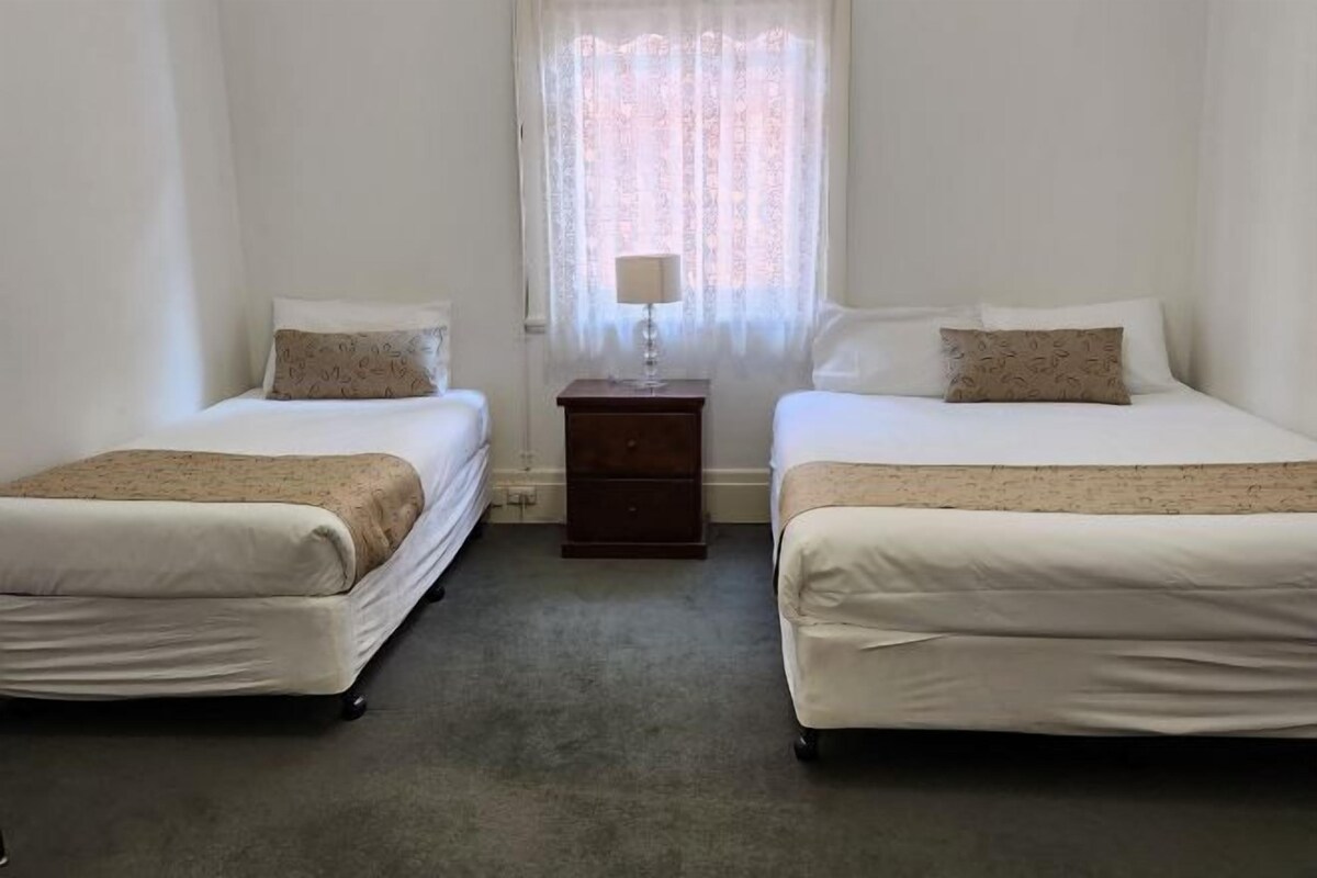Una habitación con dos camas, una mesita de noche con lámpara y una ventana con cortinas translúcidas.