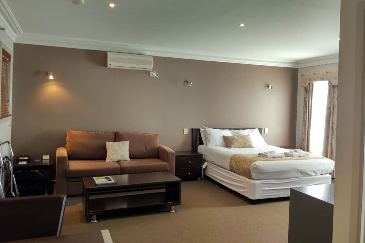 A hotel room with a brown sofa, a bed with white bedding, a bedside table, and a small desk with a chair.