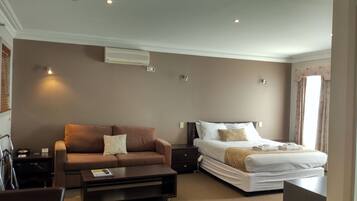 A hotel room with a brown sofa, a bed with white bedding, a bedside table, and a small desk with a chair.