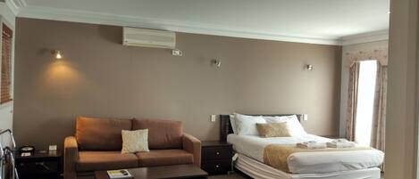 A hotel room with a brown sofa, a bed with white bedding, a bedside table, and a small desk with a chair.