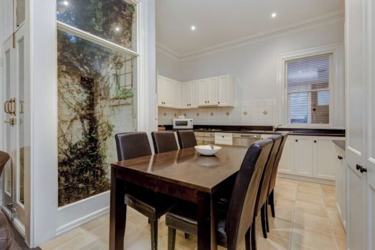 A modern kitchen with white cabinets, a dining table with chairs, and a large window with a view of trees.