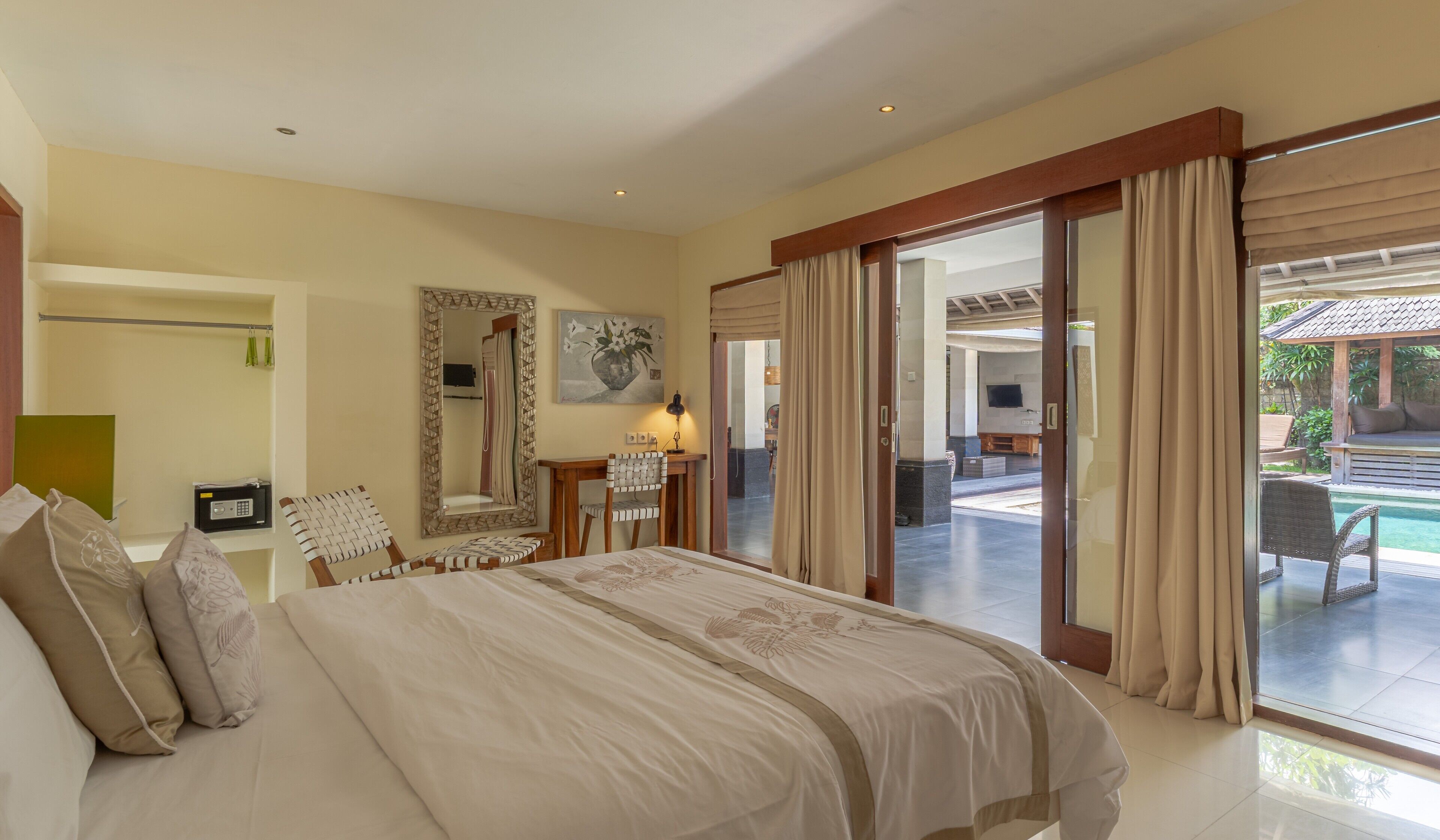 A bedroom with a large bed, a desk, a chair, a mirror, and a view of an outdoor pool area.