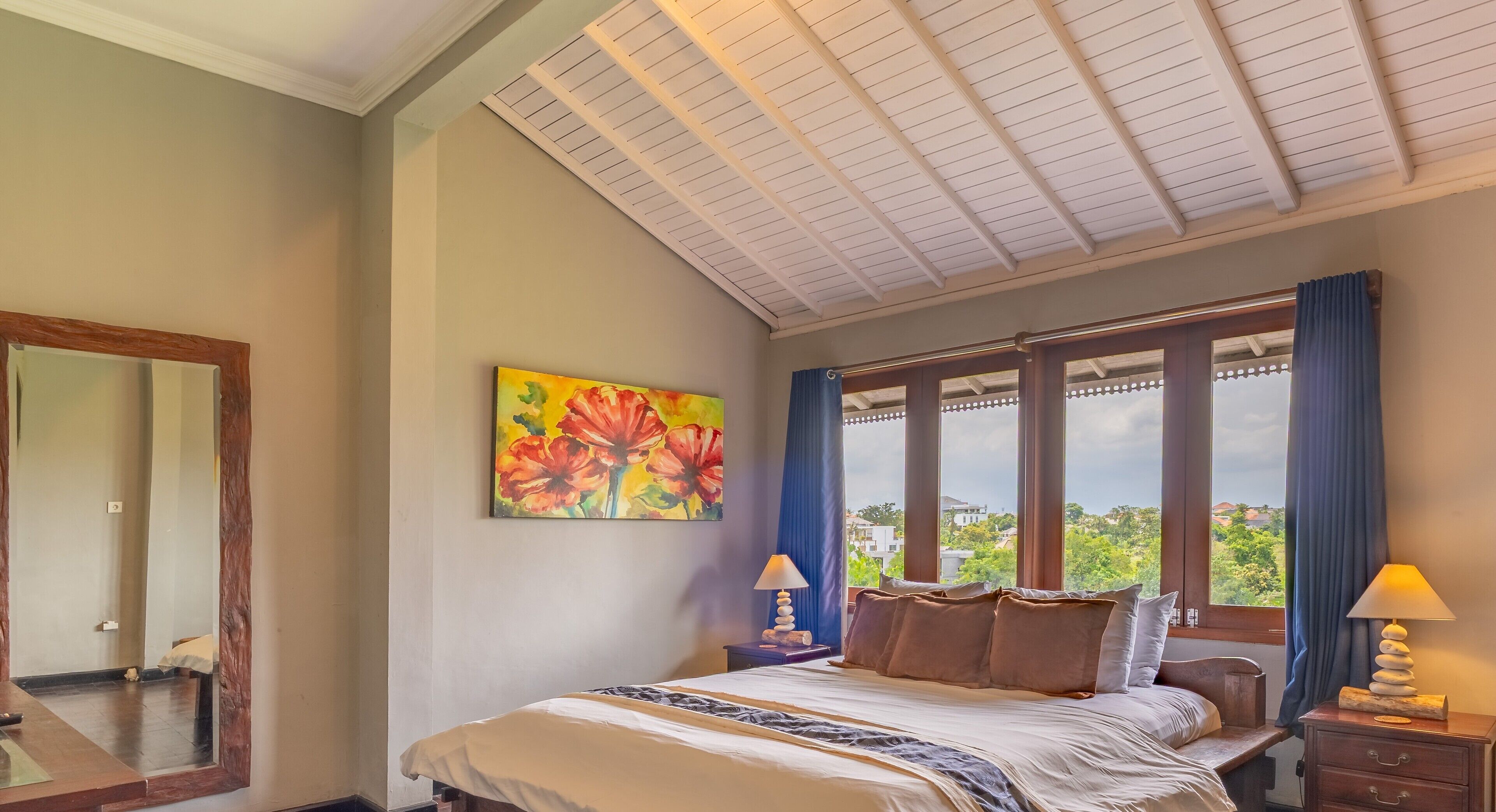 Un dormitorio con una cama grande, un cuadro de flores, una ventana con cortinas y una mesita de noche con lámpara.