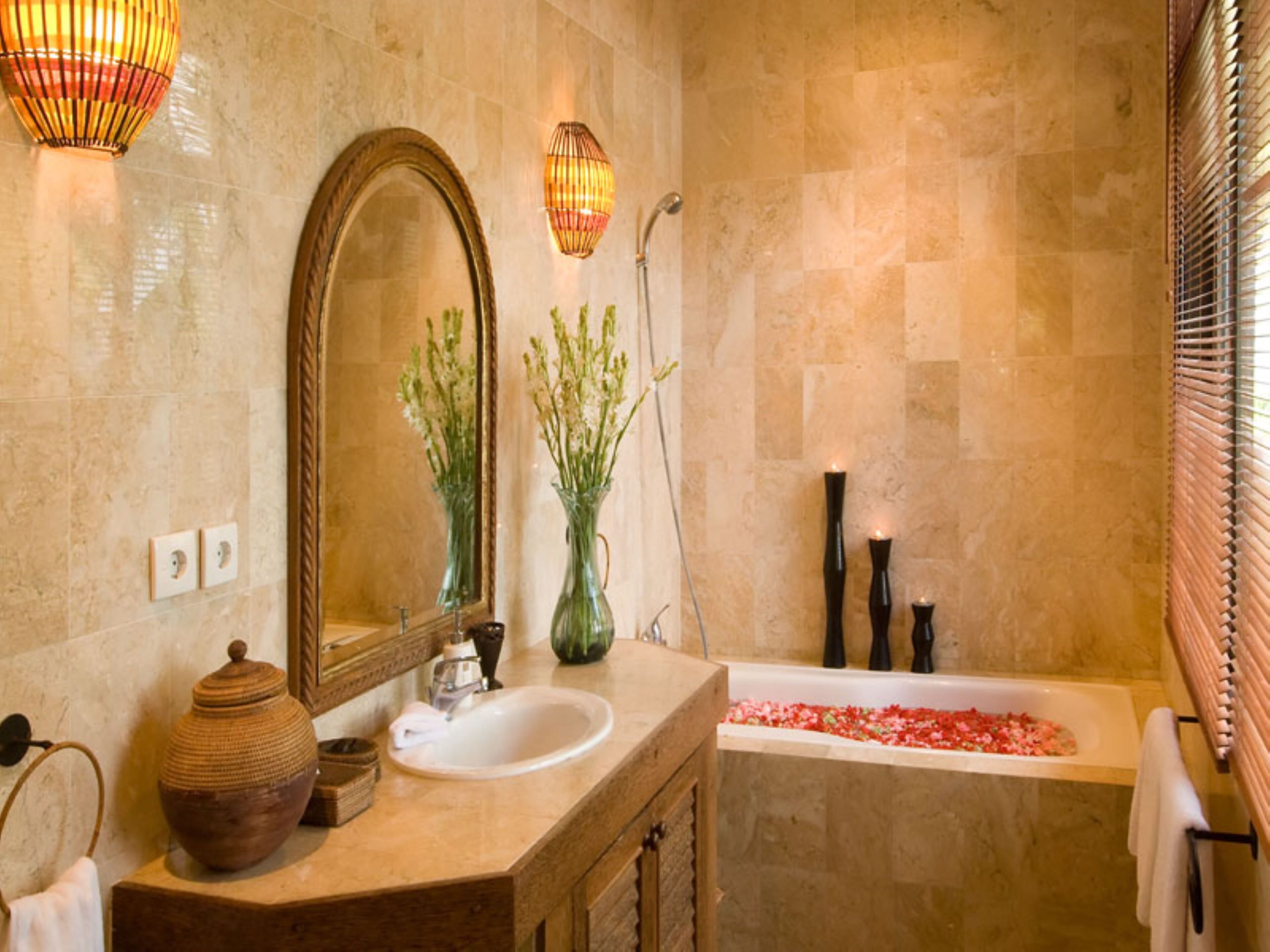 Une salle de bain avec un grand miroir, deux vases avec des plantes, une baignoire remplie de pétales de roses et un meuble-lavabo en bois.