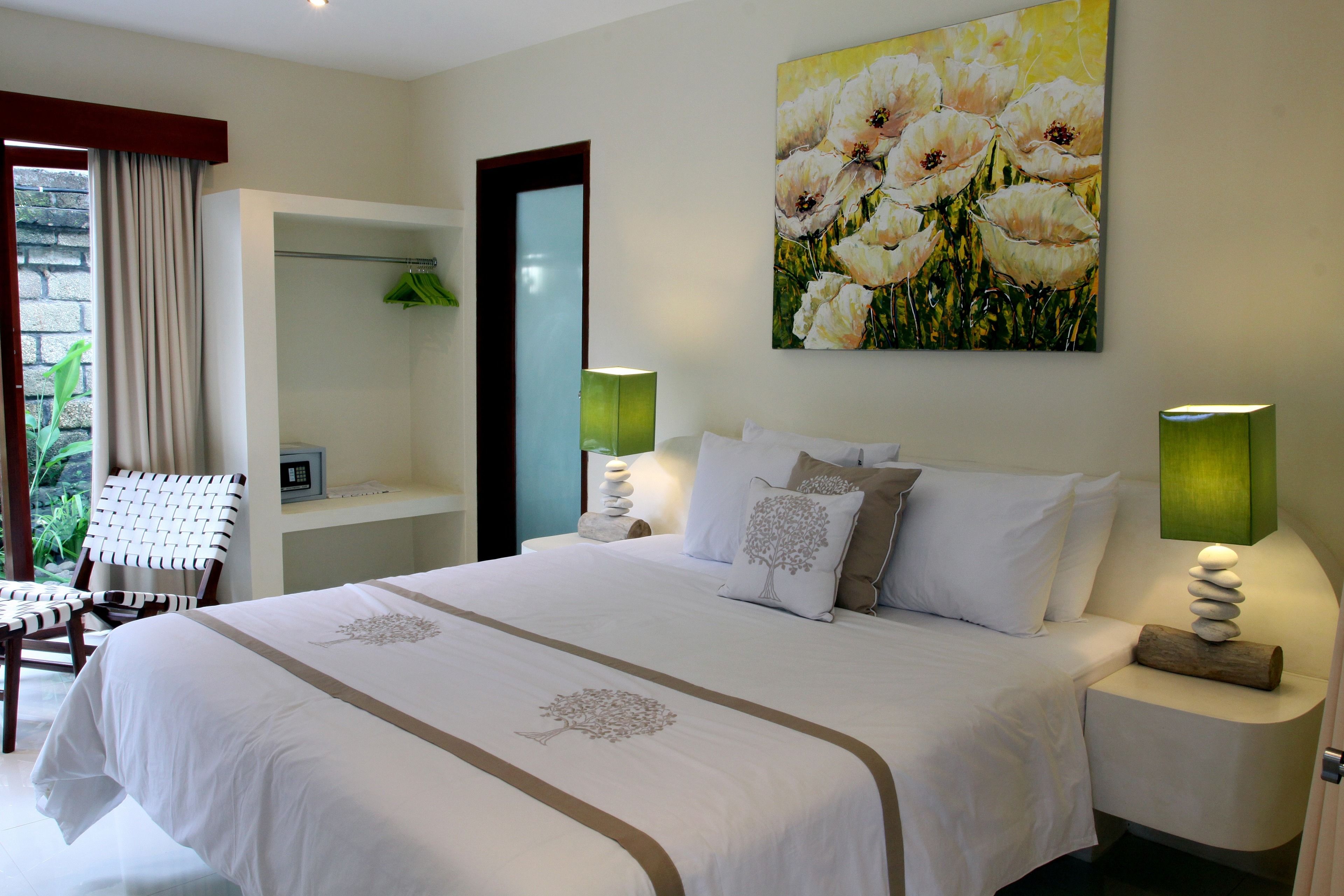 A hotel room with a large bed, two bedside tables with lamps, a painting of flowers on the wall, a chair, and a view of greenery outside.