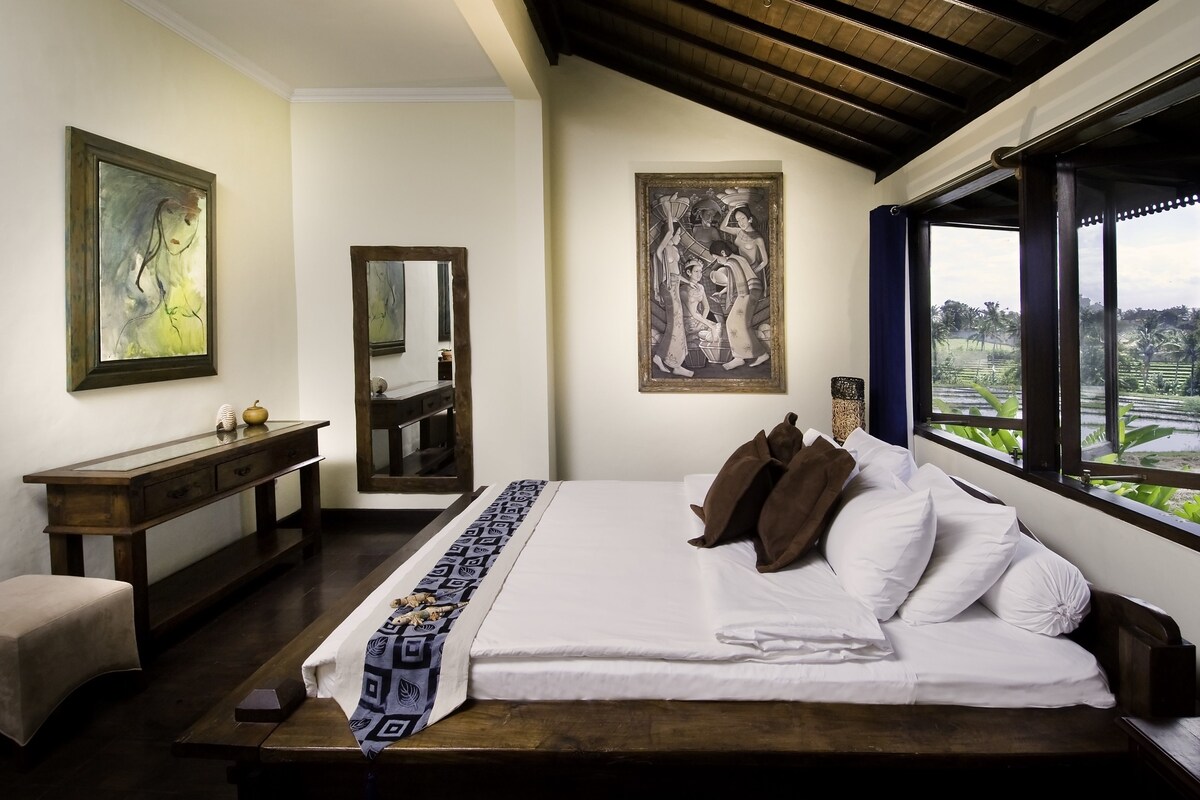 Un dormitorio con una cama grande, una mesita de noche de madera, un aparador y un espejo. Hay dos cuadros en las paredes y una ventana con vistas a la vegetación.