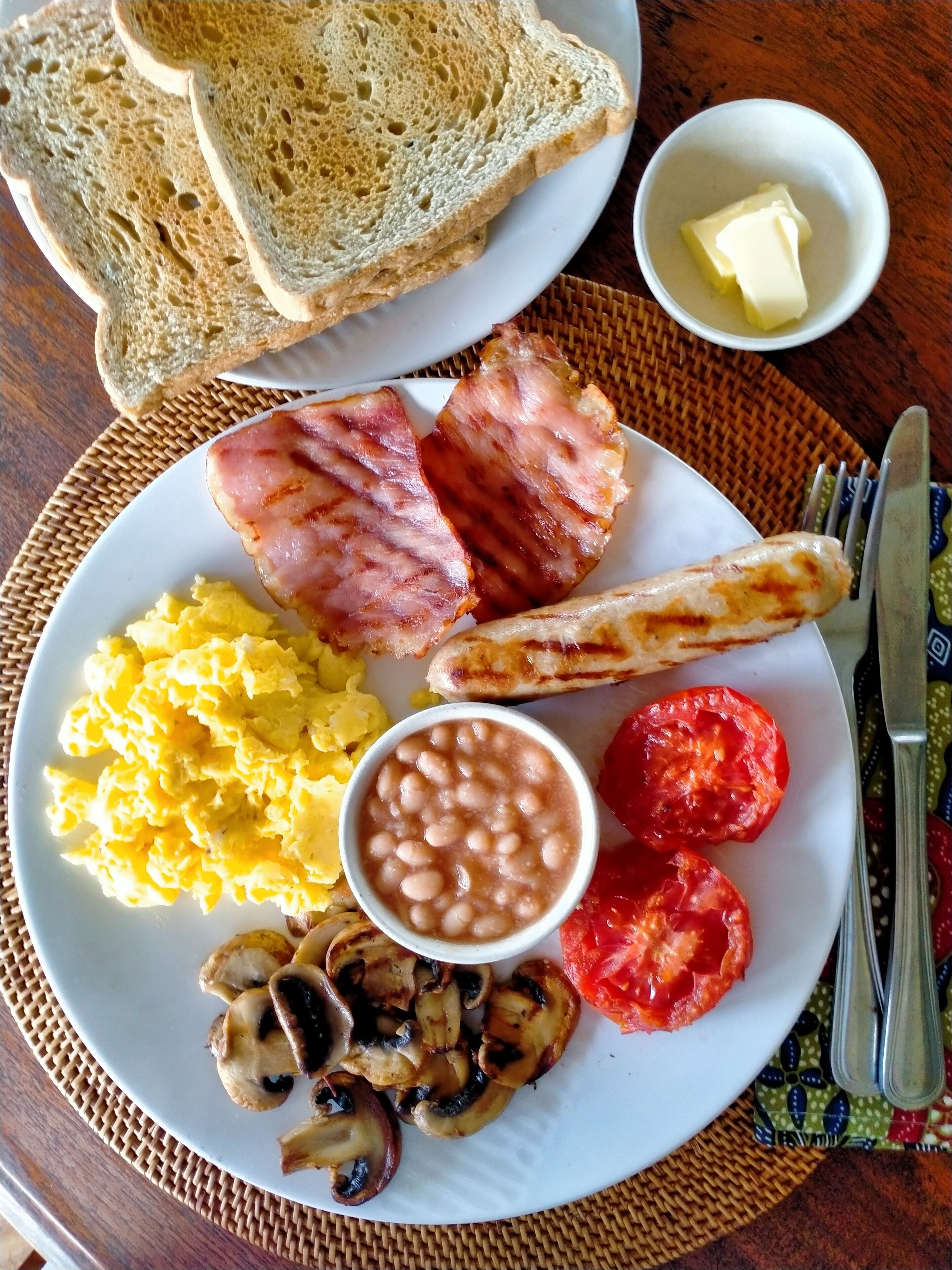 A plate with scrambled eggs, bacon, grilled mushrooms, tomatoes, beans, toast, and butter on a table.