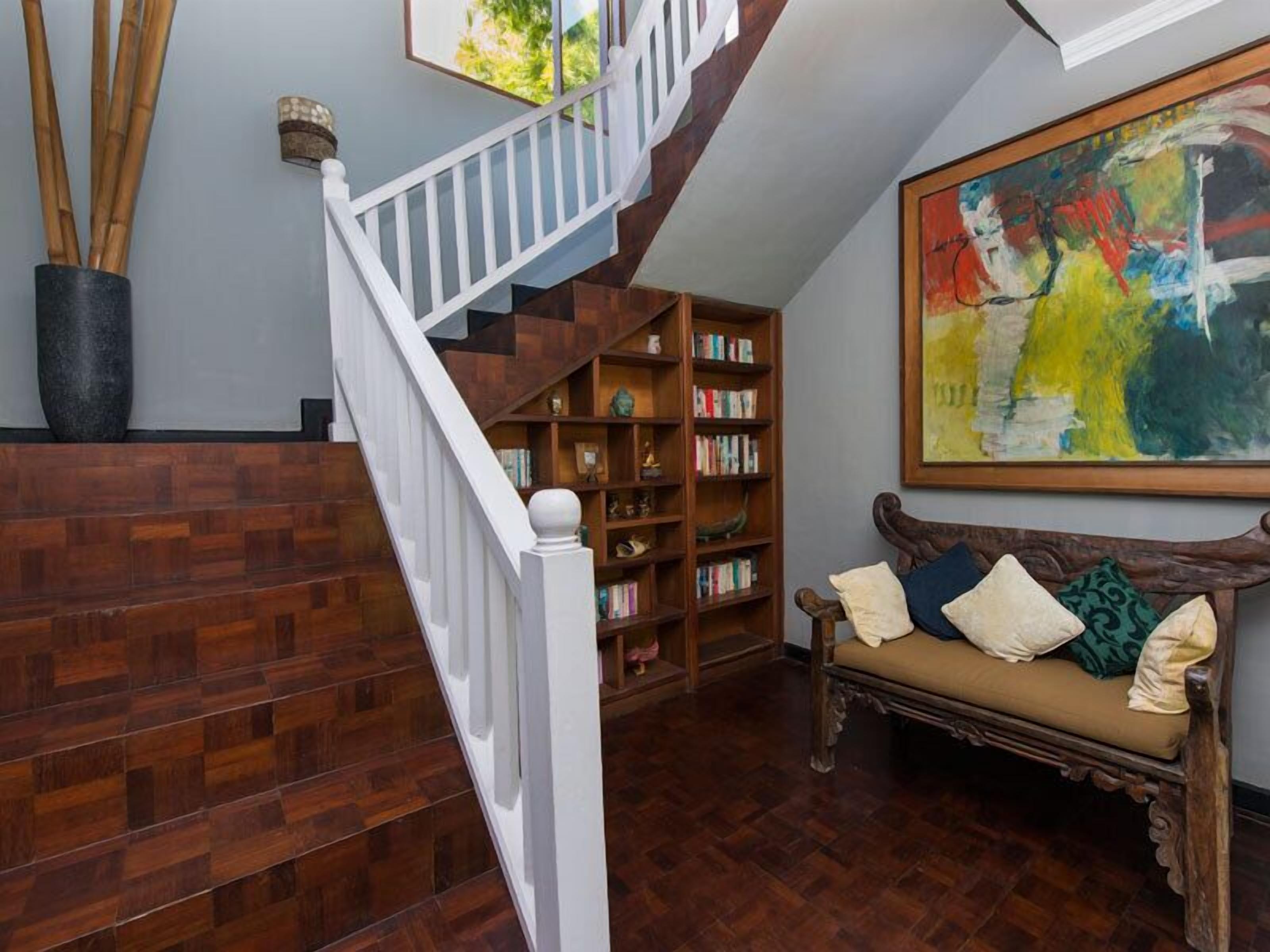 A wooden staircase with a white railing, a bookshelf filled with books, a cushioned bench with pillows, and a large abstract painting on the wall.