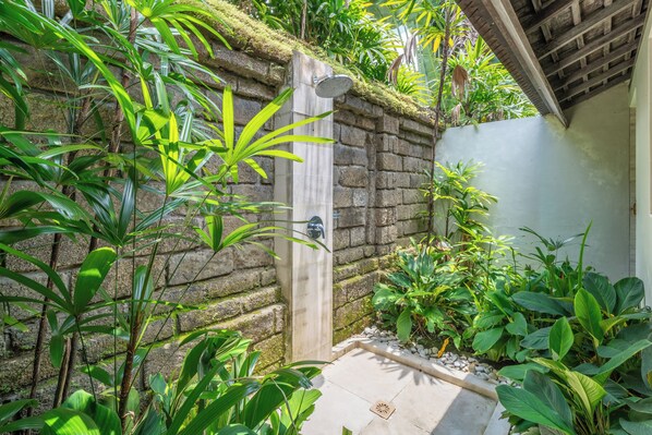 Zona de ducha con paredes de piedra, puerta de madera y abundante vegetación verde.