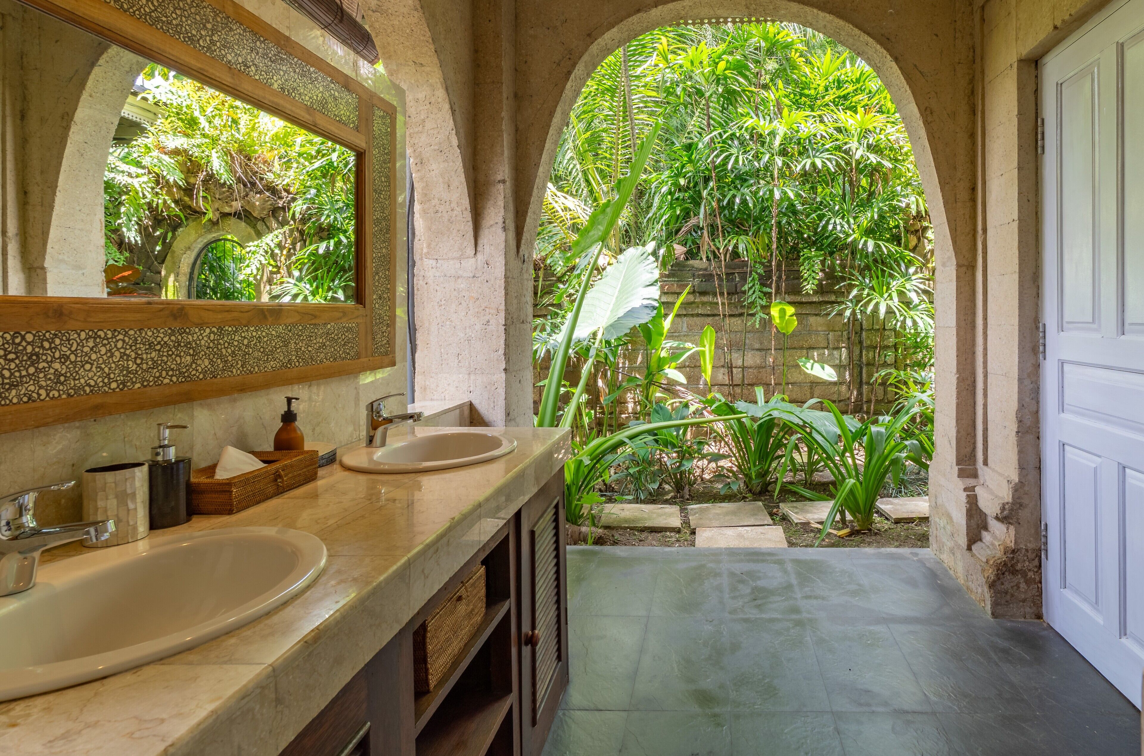 Un baño con dos lavamanos, un espejo y vistas a un jardín exuberante a través de un arco.