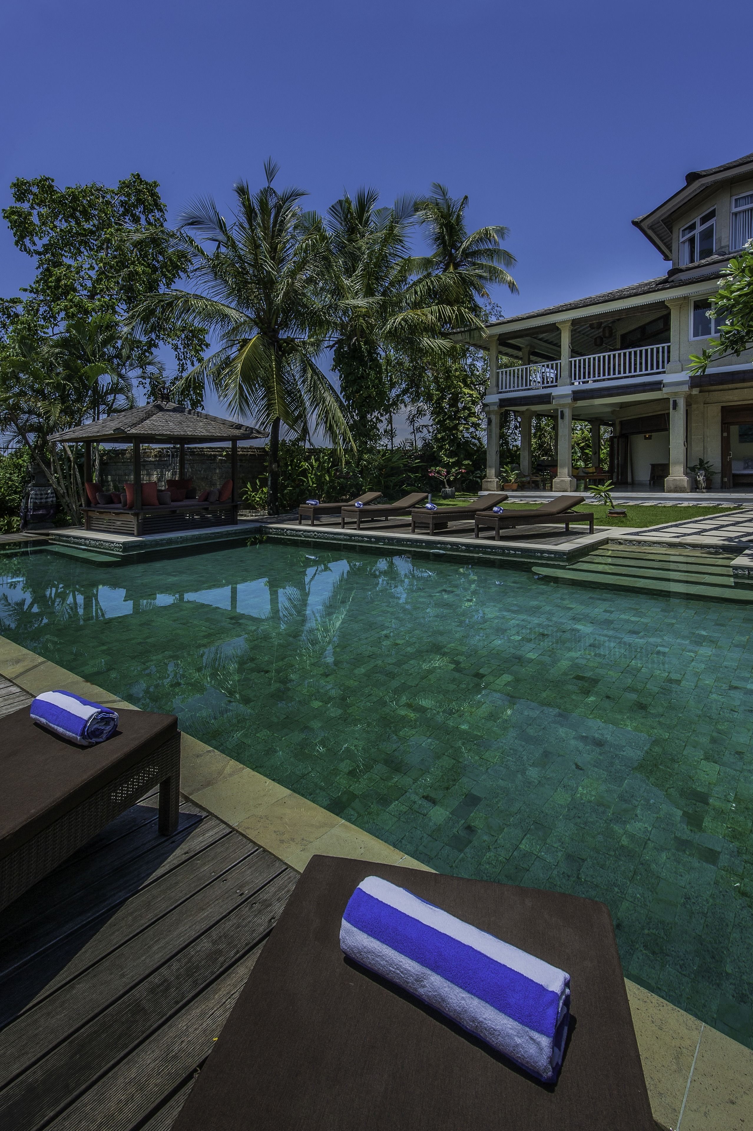 A clear pool with lounge chairs and a gazebo, surrounded by palm trees and a large building.
