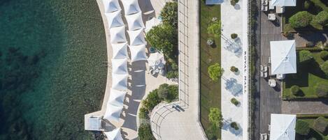An aerial view of a coastal hotel with rows of white umbrellas, a paved walkway, and a clear view of the sea.