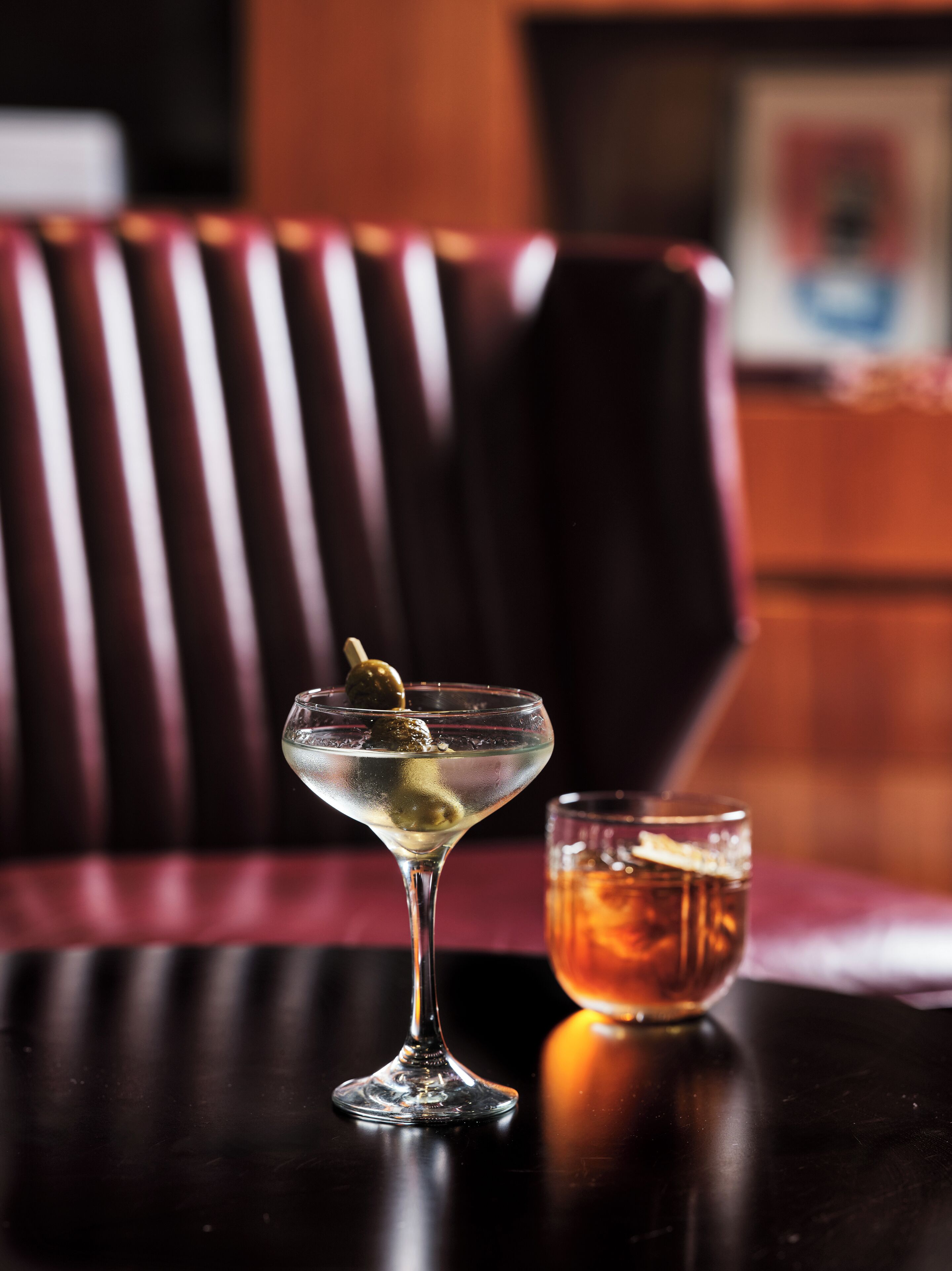A martini glass with an olive on a dark wooden table, a glass of amber-colored drink, and a striped red and black chair in the background.