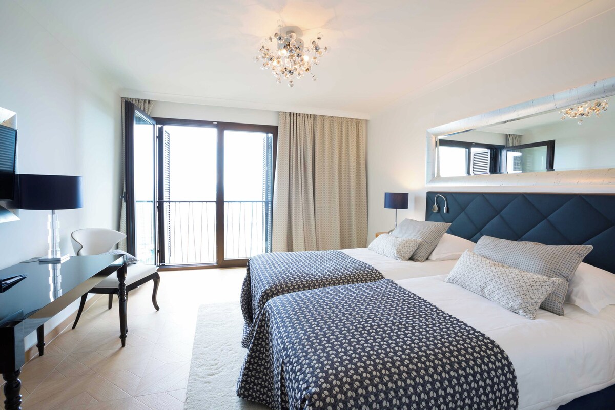 Una habitación de hotel con una cama grande, dos mesitas de noche, un escritorio y una silla. Hay un ventanal con cortinas y un candelabro.