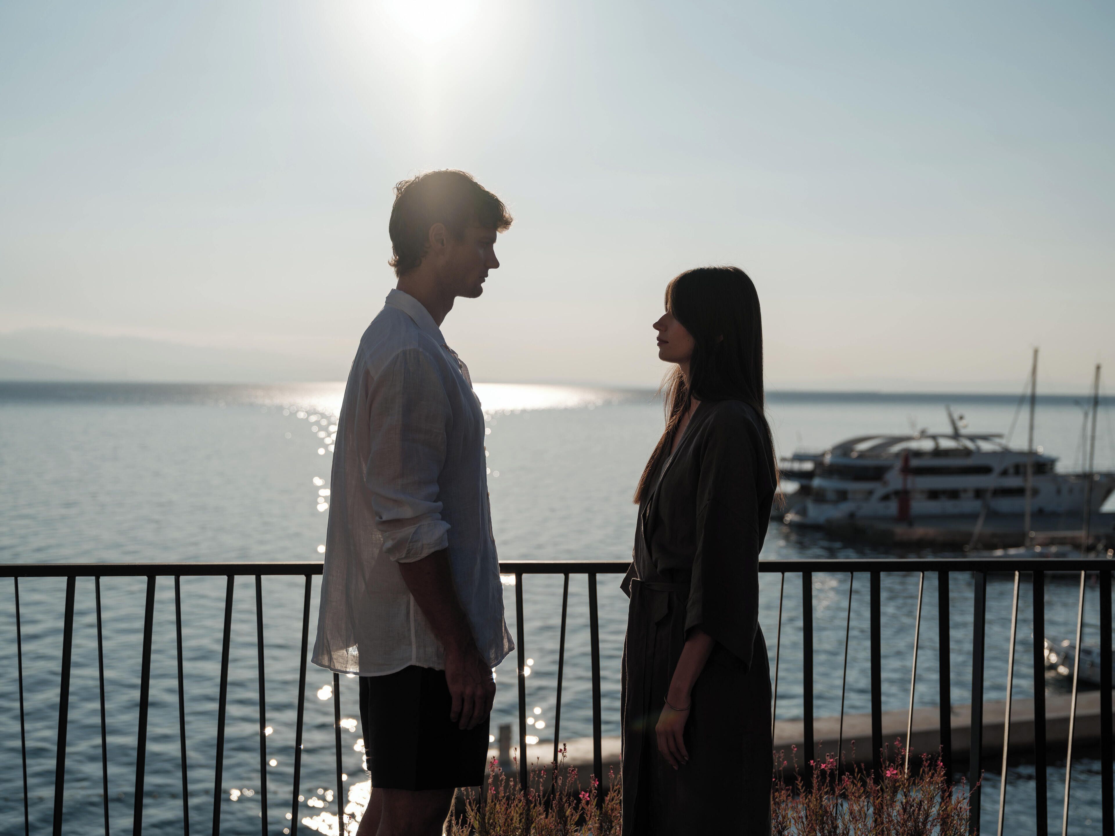 A couple standing on a balcony overlooking a body of water with boats in the background.