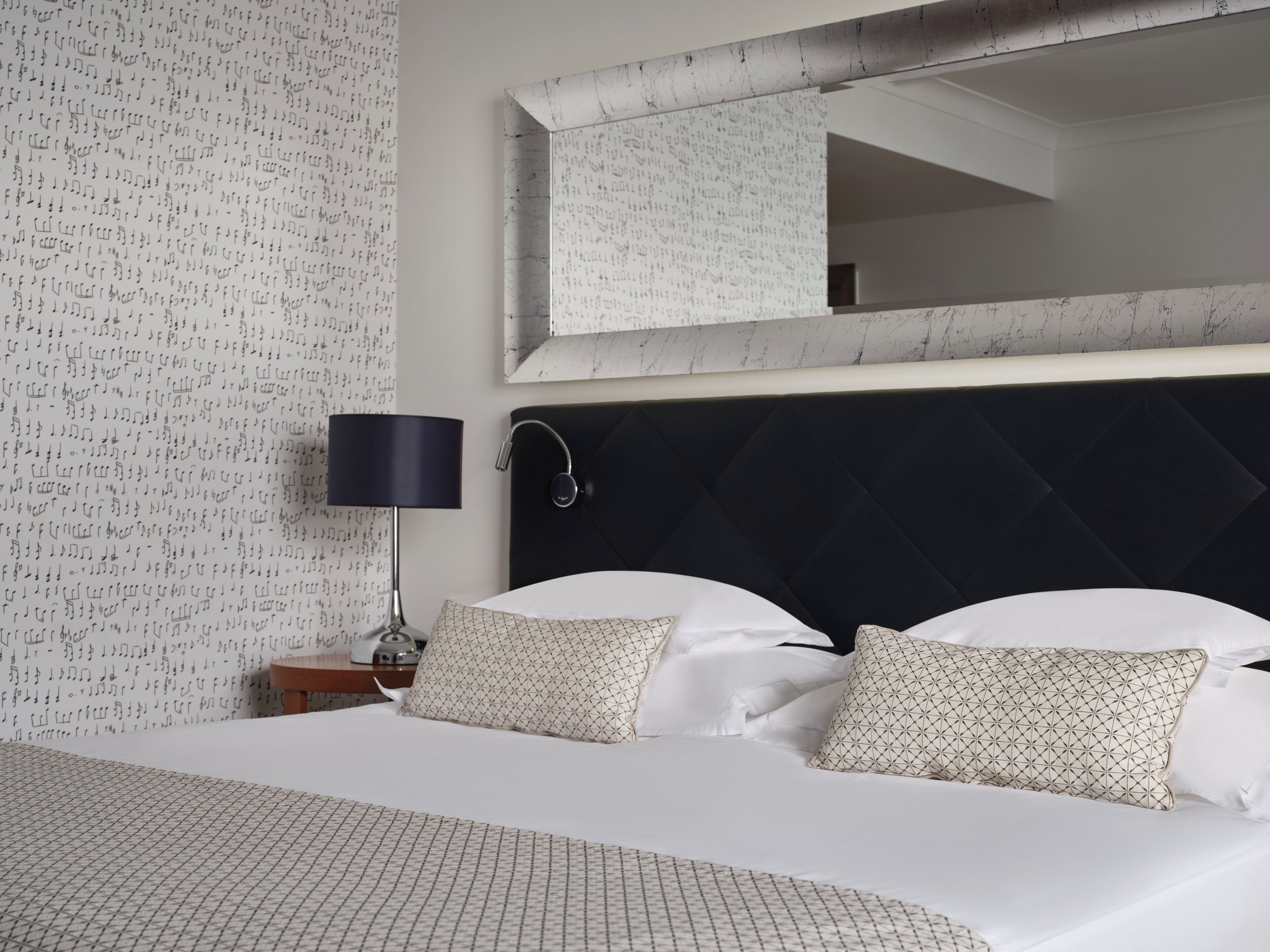 A hotel room with a bed, a nightstand, a lamp, and a wall with a musical note pattern.