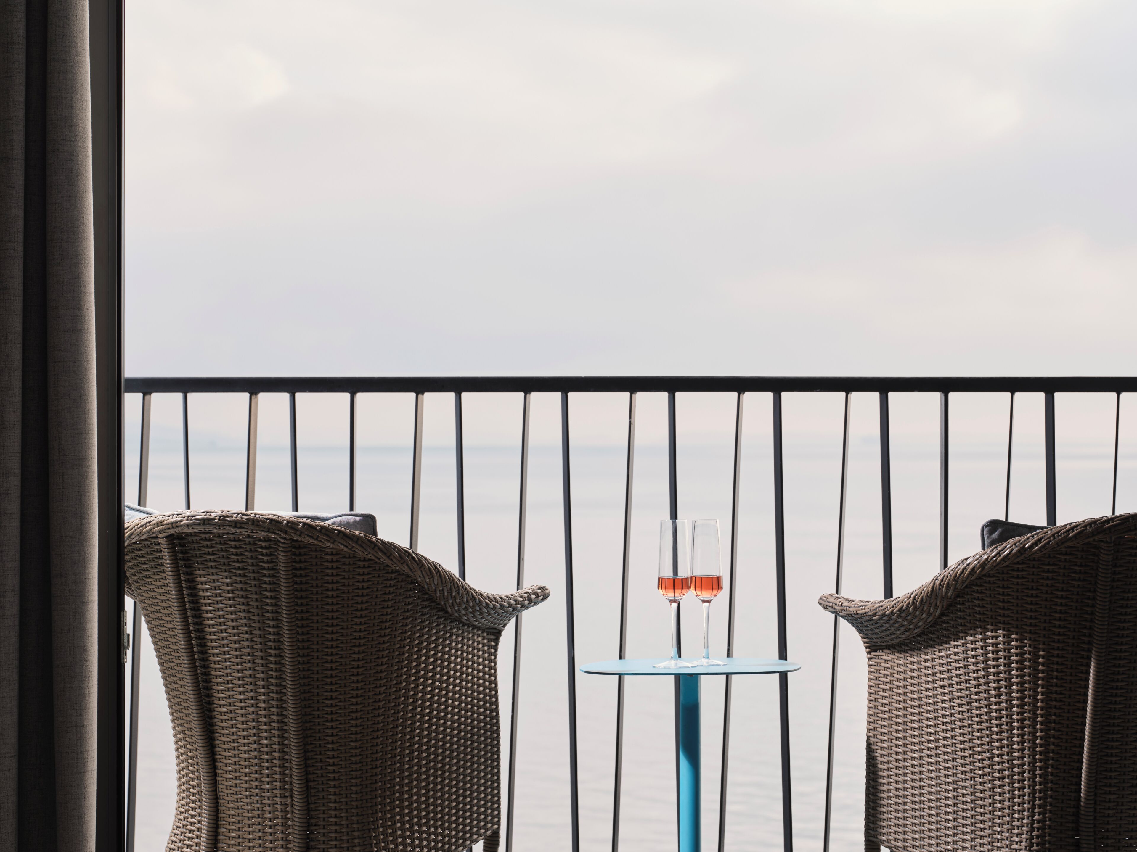 Een balkon met rieten stoelen, een klein tafeltje en twee glazen wijn.