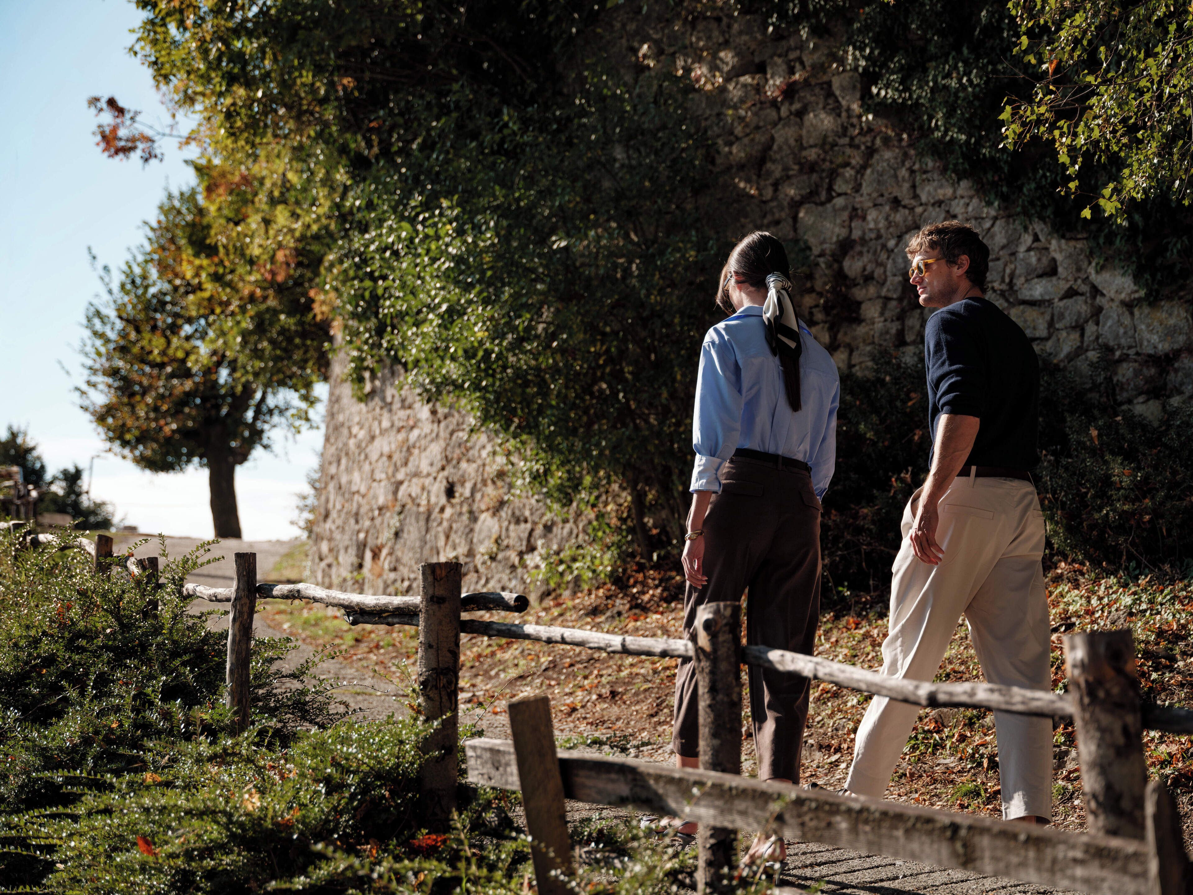 Two people walking on a stone pathway with a rustic wooden fence, surrounded by greenery and a stone wall.