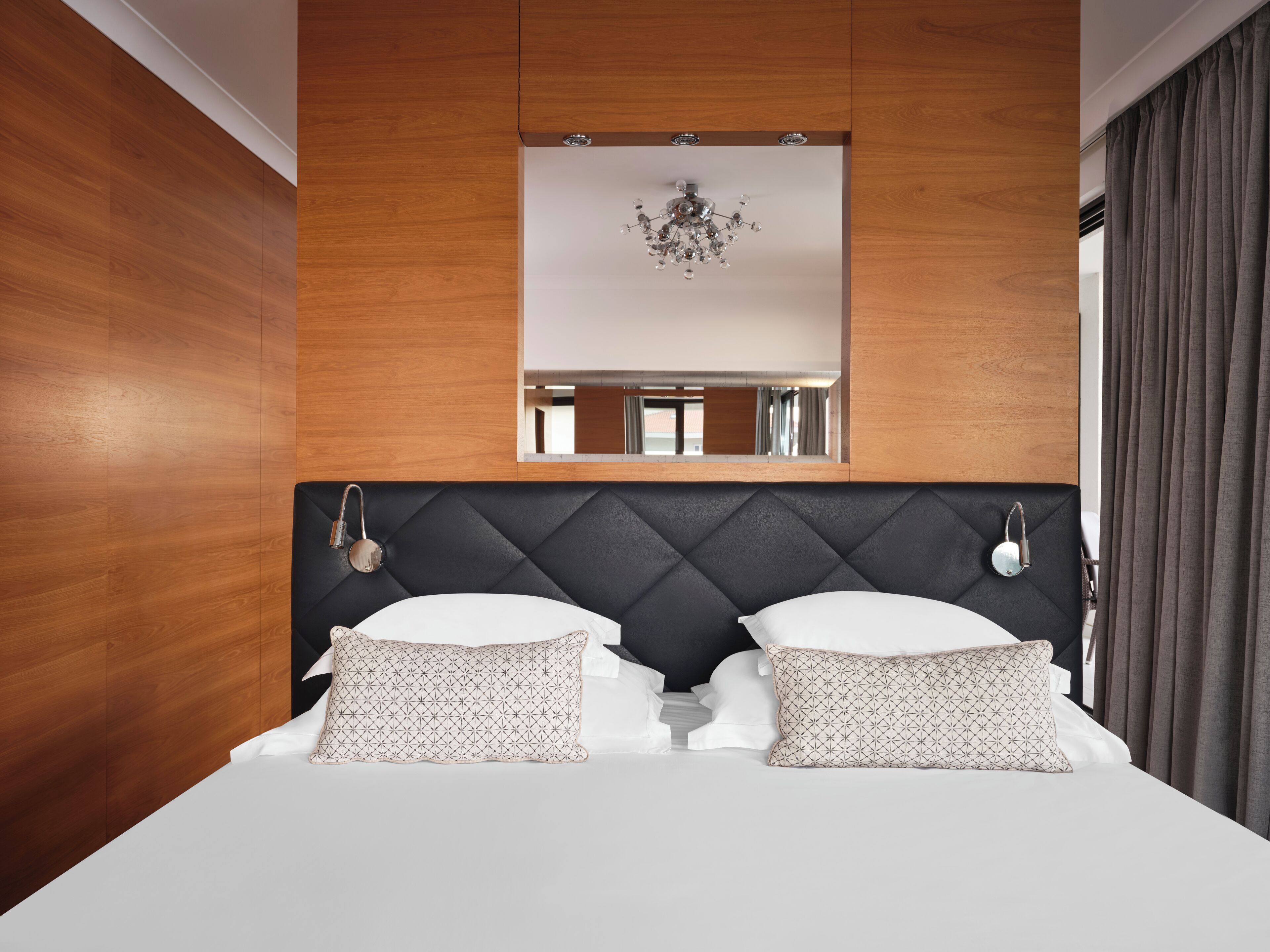Une chambre d’hôtel moderne avec un mur lambrissé en bois, une tête de lit noire à capitons, un lustre et un grand miroir au-dessus du lit.
