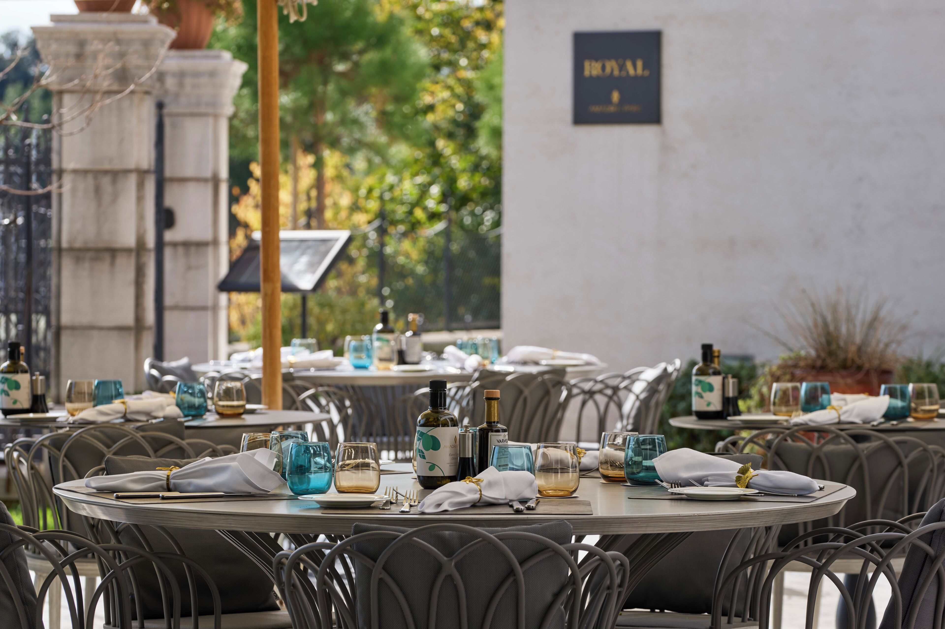 A restaurant setting with round tables, chairs, and neatly set dining tables with glasses, bottles, and napkins.