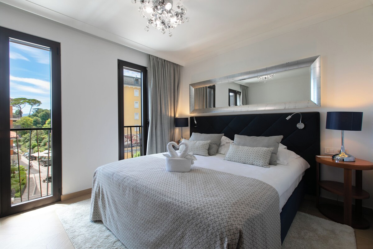 A bedroom with a large bed, a nightstand, a mirror, and a view of the outdoors.