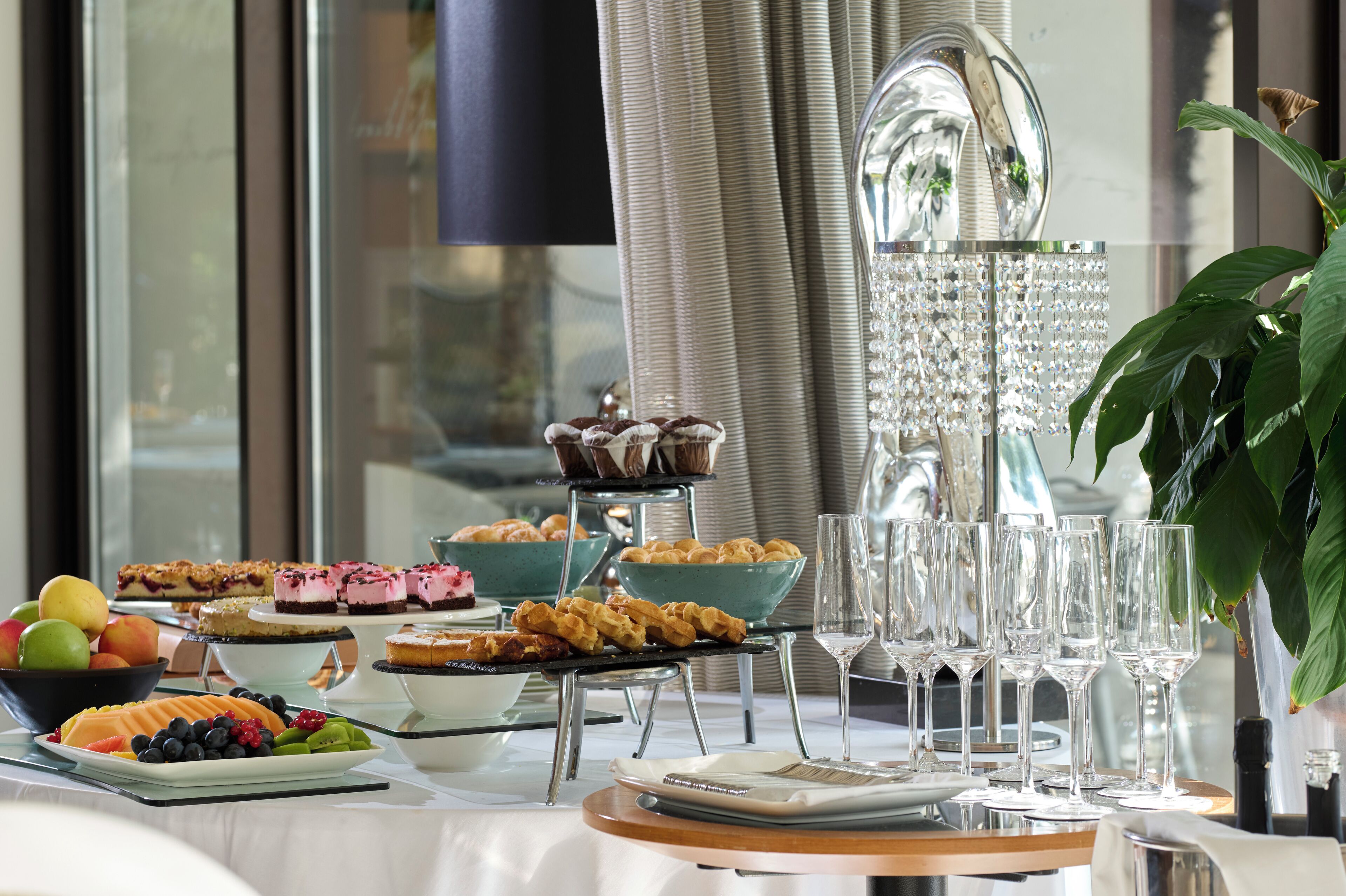 A buffet table with various desserts and fruits, glassware, and a potted plant.