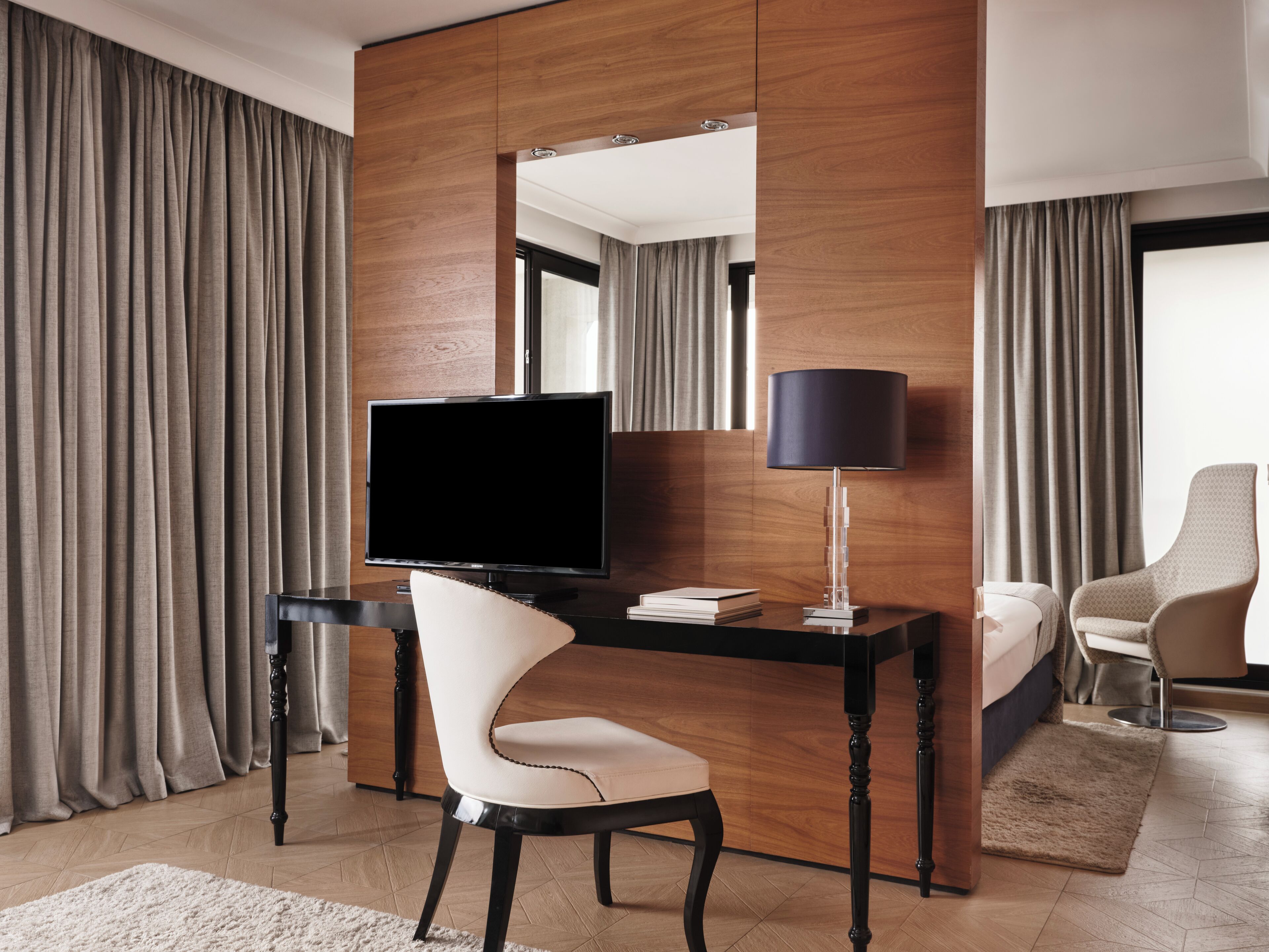 A hotel room with a wooden desk, a television, a lamp, a chair, a bed, and curtains.