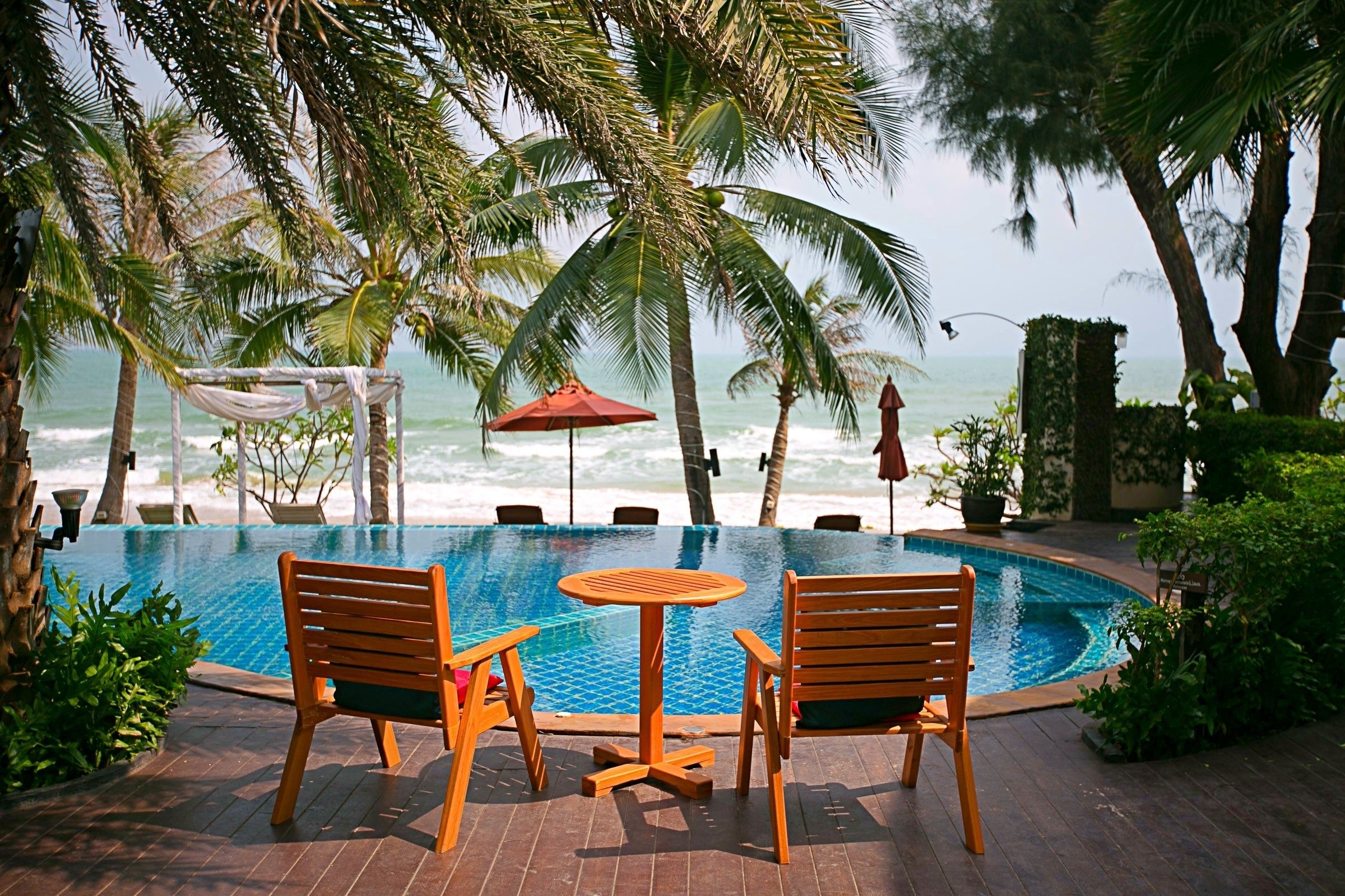 A pool area with wooden chairs and a table, surrounded by palm trees and overlooking the ocean. - iL Mare Resort (Pranburi)