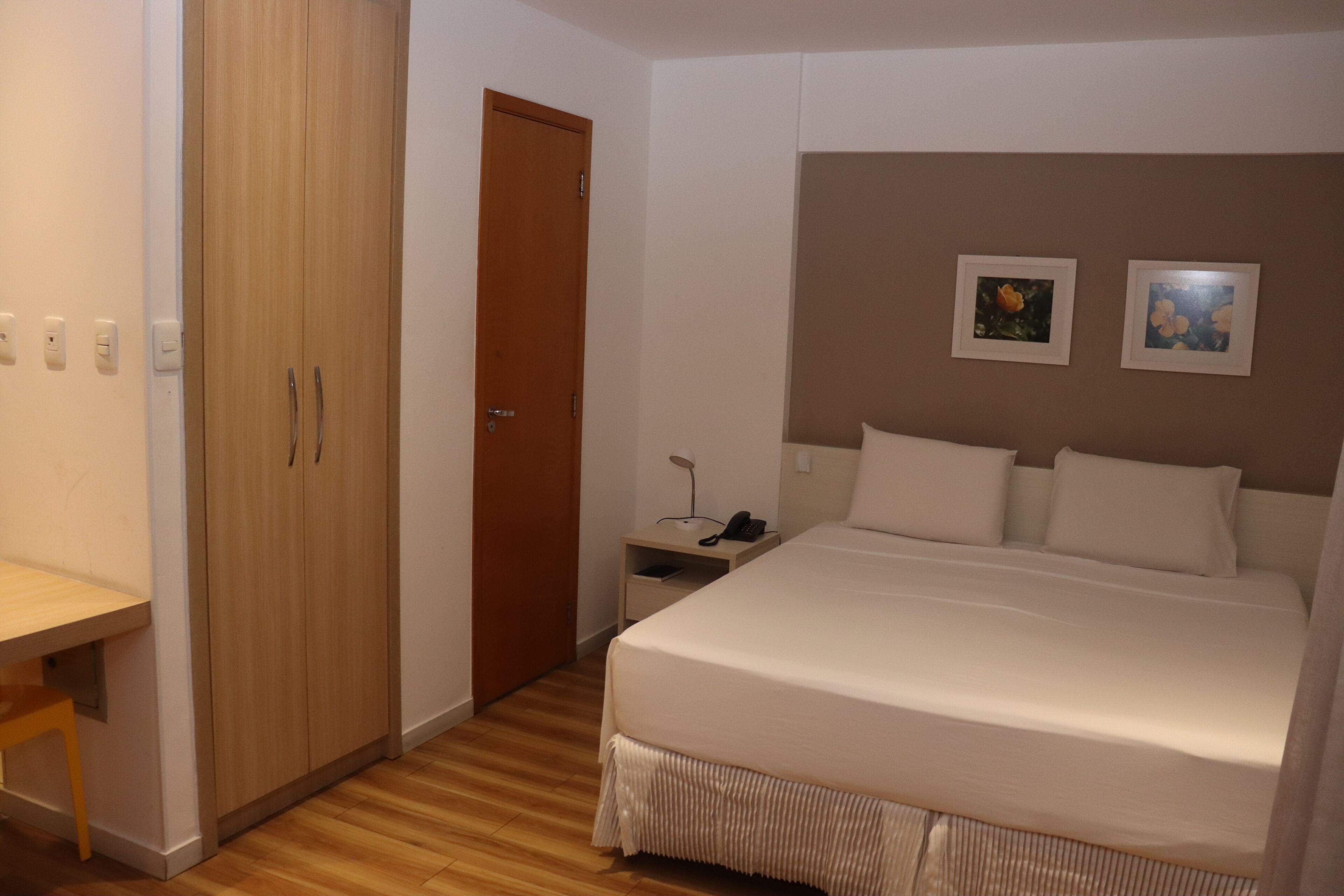 A bedroom with a wooden wardrobe, a bedside table with a lamp, and two framed pictures on the wall.