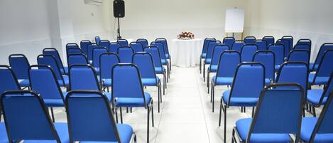 Une salle de conférence avec des rangées de chaises bleues, une table blanche agrémentée d’un arrangement floral et un tableau blanc.