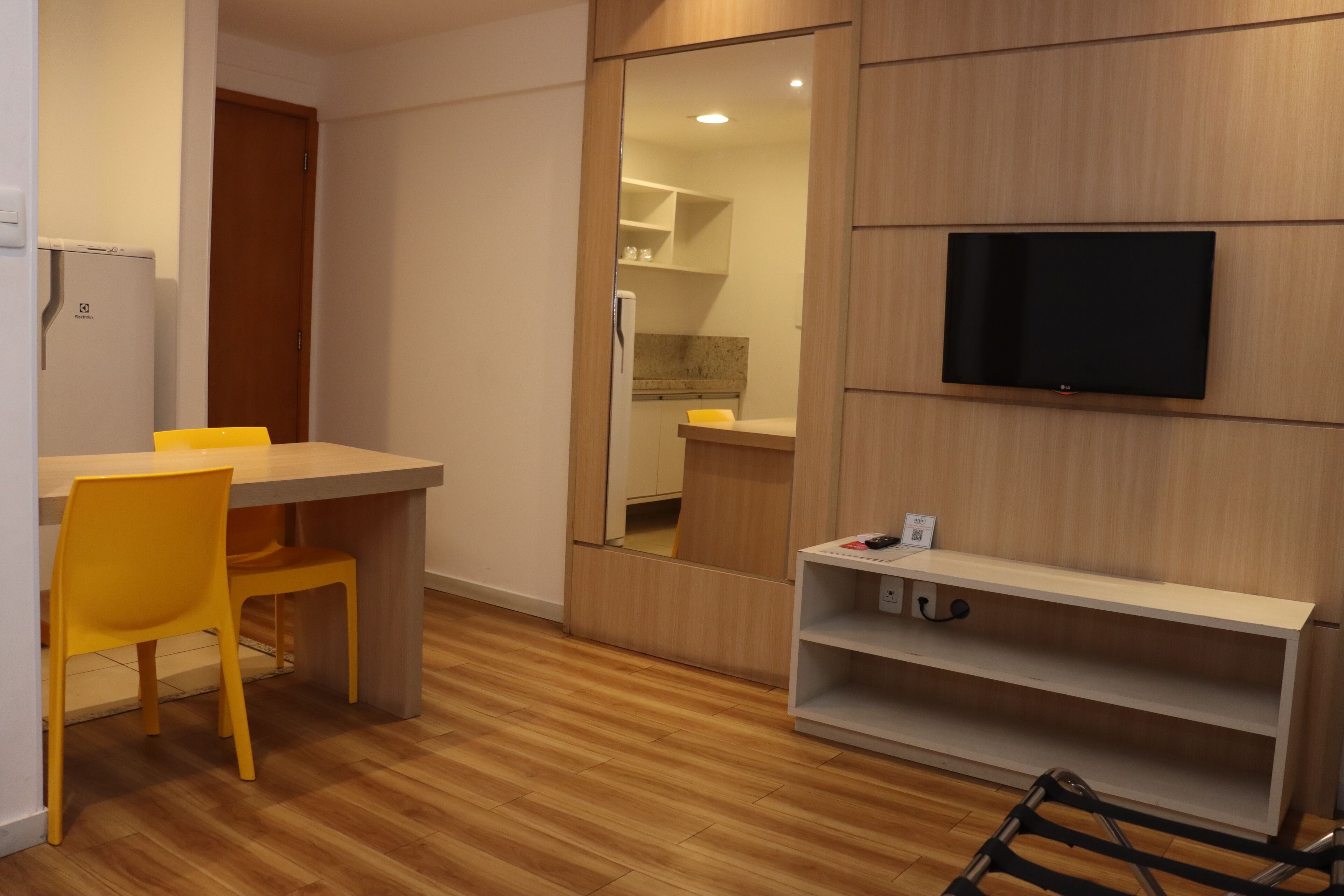 Ein modernes Zimmer mit Holzboden, einem Esstisch mit gelben Stühlen, einem an der Wand befestigten Fernseher und einem Spiegel, der den Küchenbereich reflektiert.