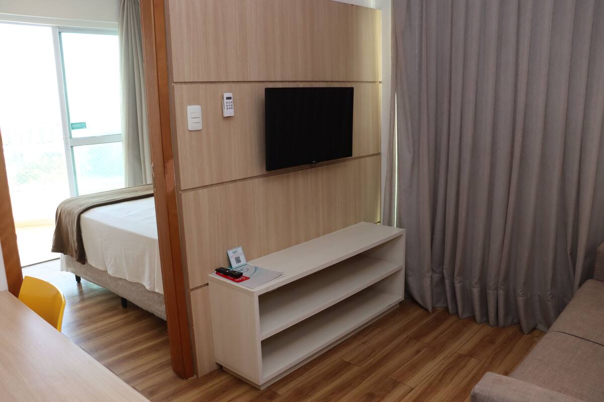 Een moderne hotelkamer met een bed, een tv die aan een houten paneel is bevestigd, een klein bureau met een afstandsbediening en een bank.