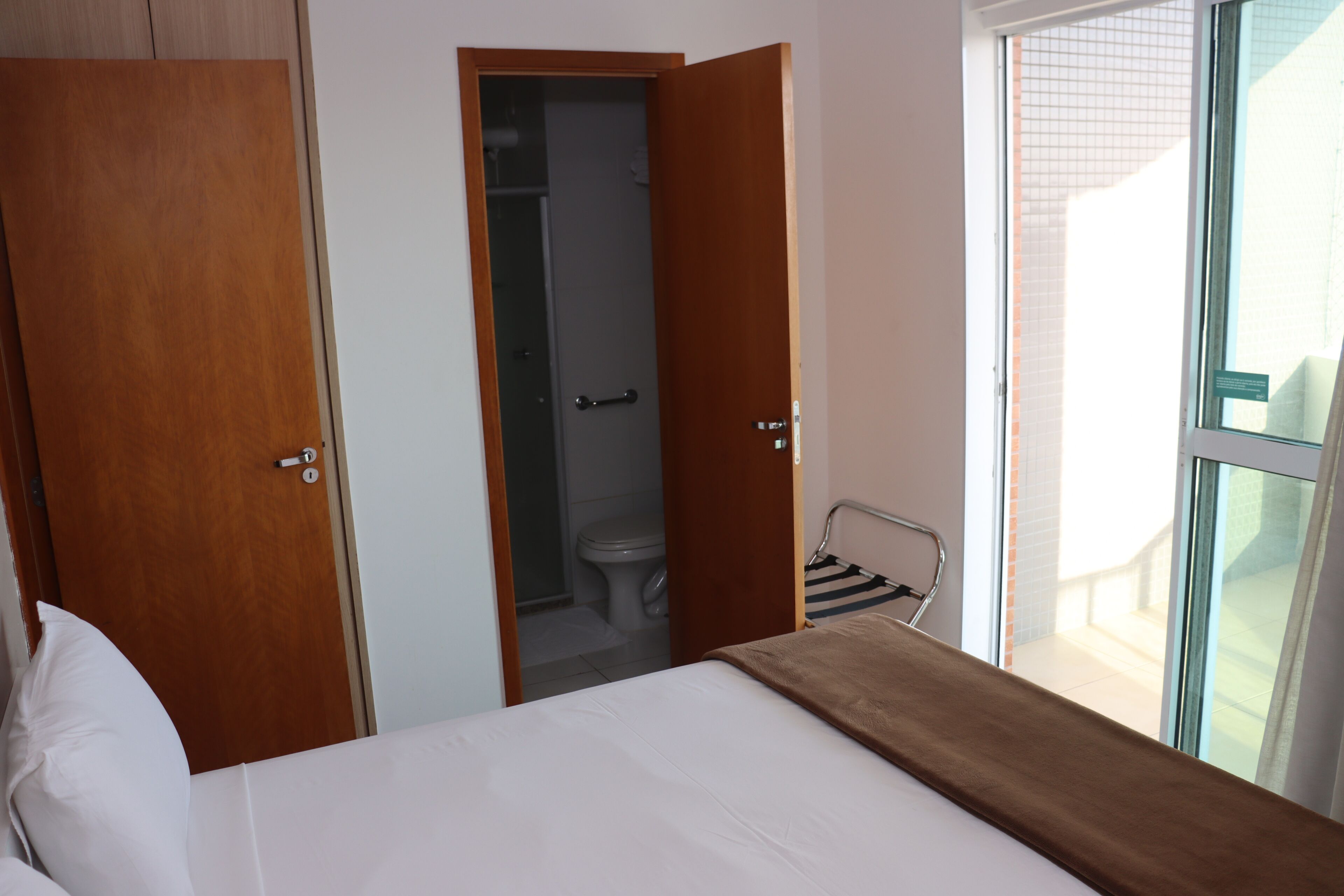 A bedroom with a bed, a wooden wardrobe, a bathroom visible through an open door, and a window with a view of the outside.