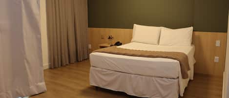 Una habitación de hotel con cama, cortinas, mesita de noche y suelo de madera.
