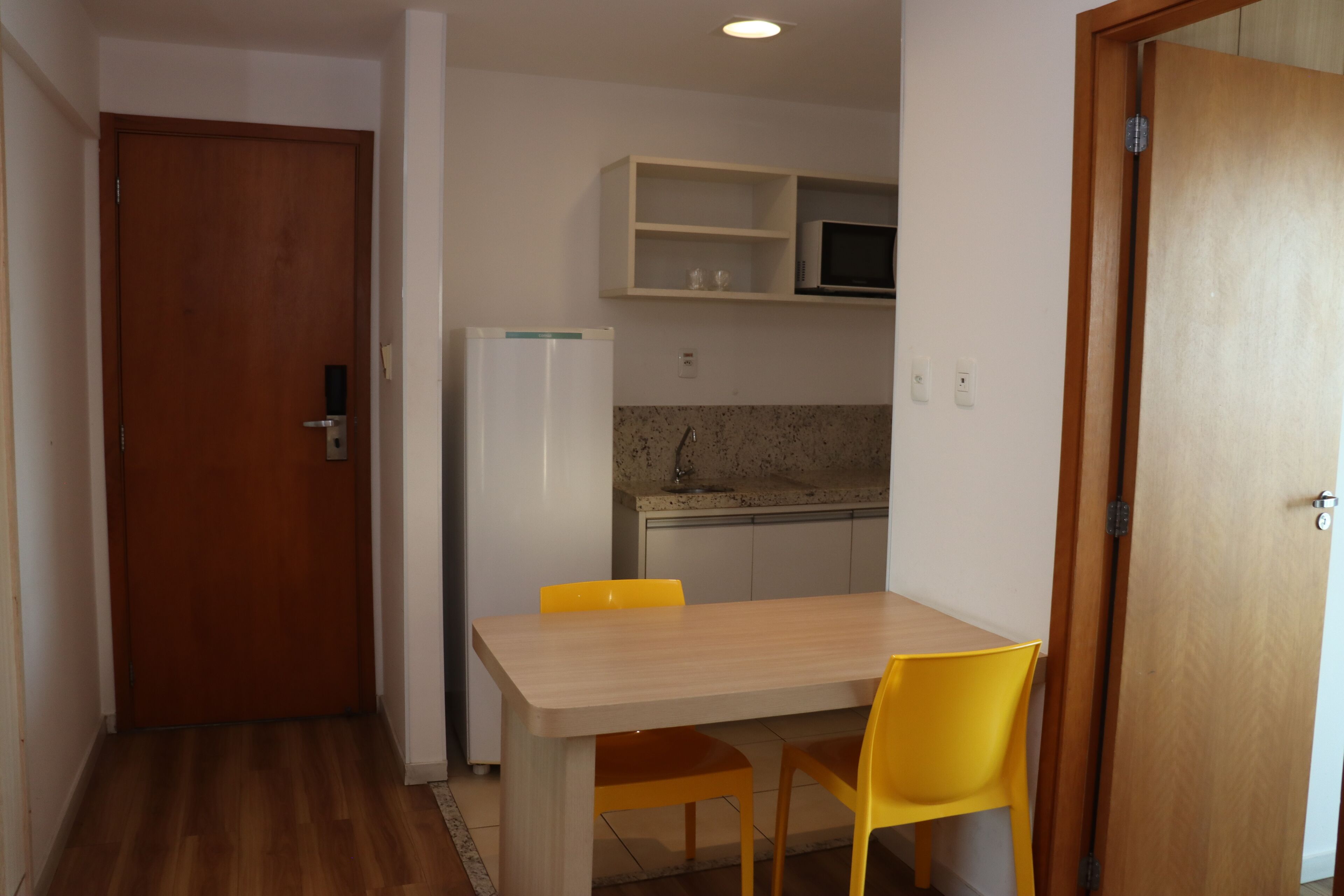 A compact kitchen with a microwave, a refrigerator, and a sink. There is a wooden door on the left and a wooden-framed door on the right.