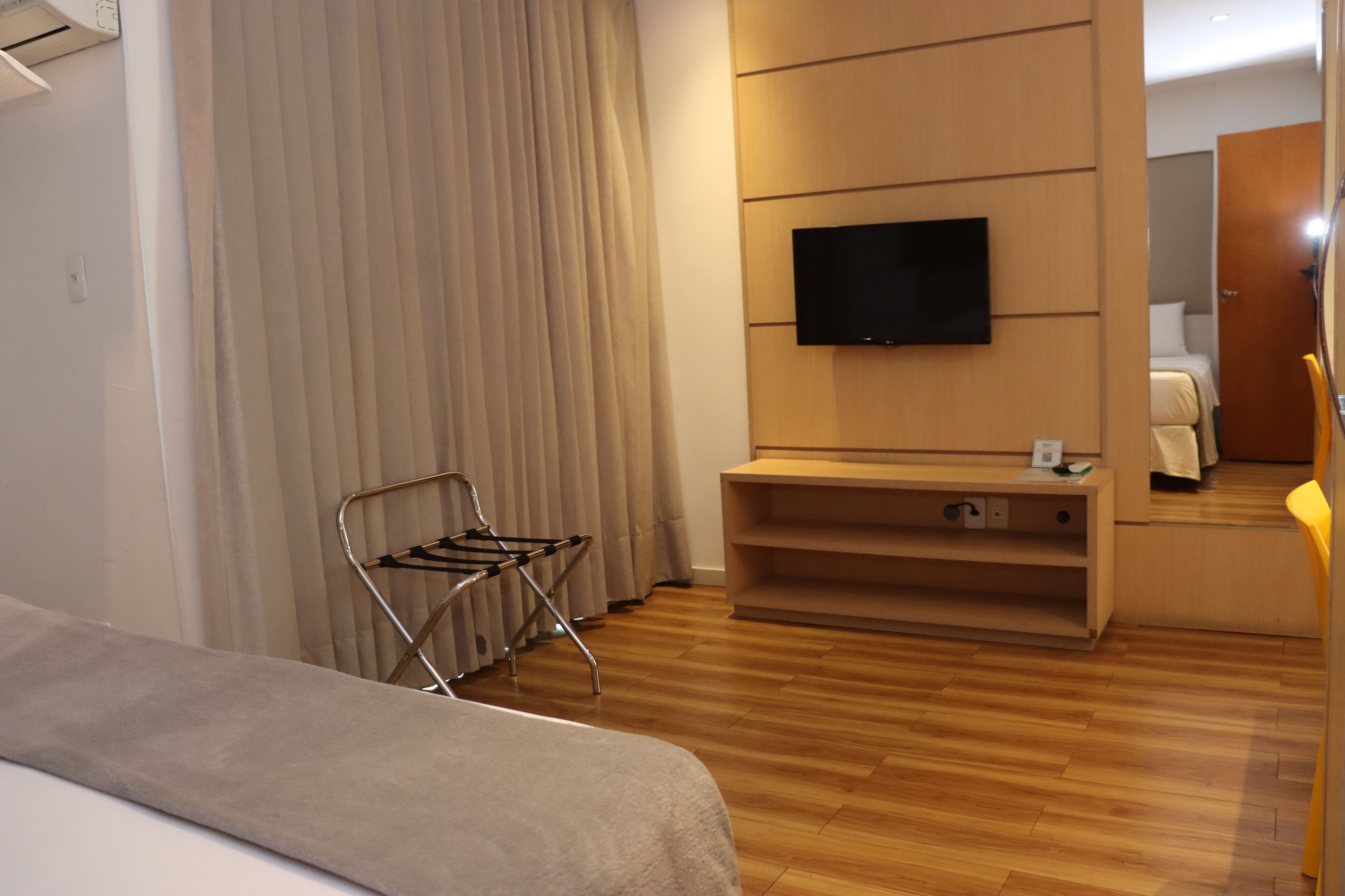Habitación de hotel con cama, televisor montado en la pared, piso de madera y ventana con cortinas.