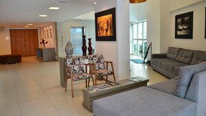 A modern living room with a grey sofa, a coffee table with magazines, patterned chairs, a wooden side table with vases, and artwork on the walls. - Único Apart Hotel (Feira de Santana)