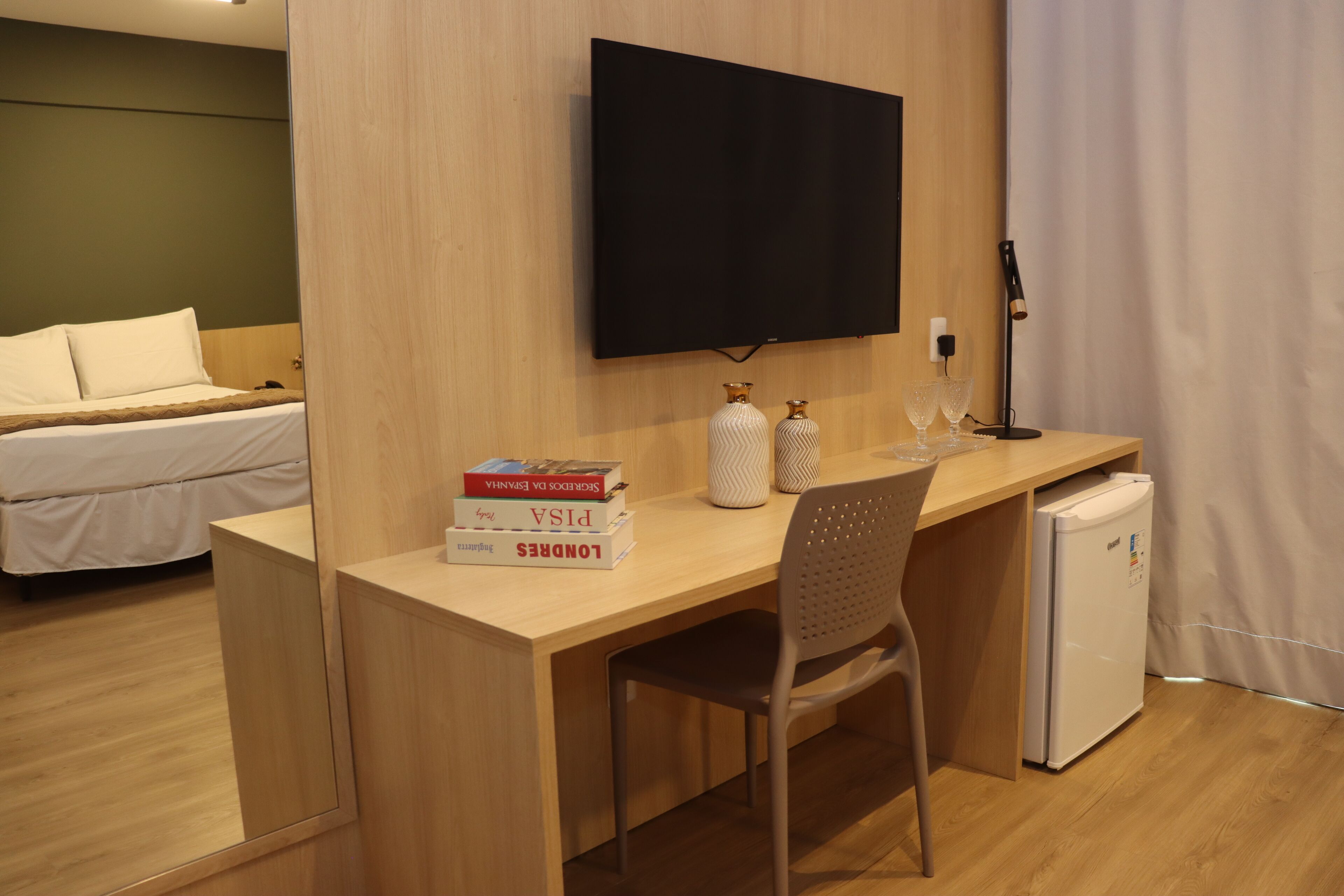 A hotel room with a wooden desk, a flat-screen TV mounted on the wall, a small refrigerator, a bed with white bedding, and a bedside table with a lamp and two decorative vases.