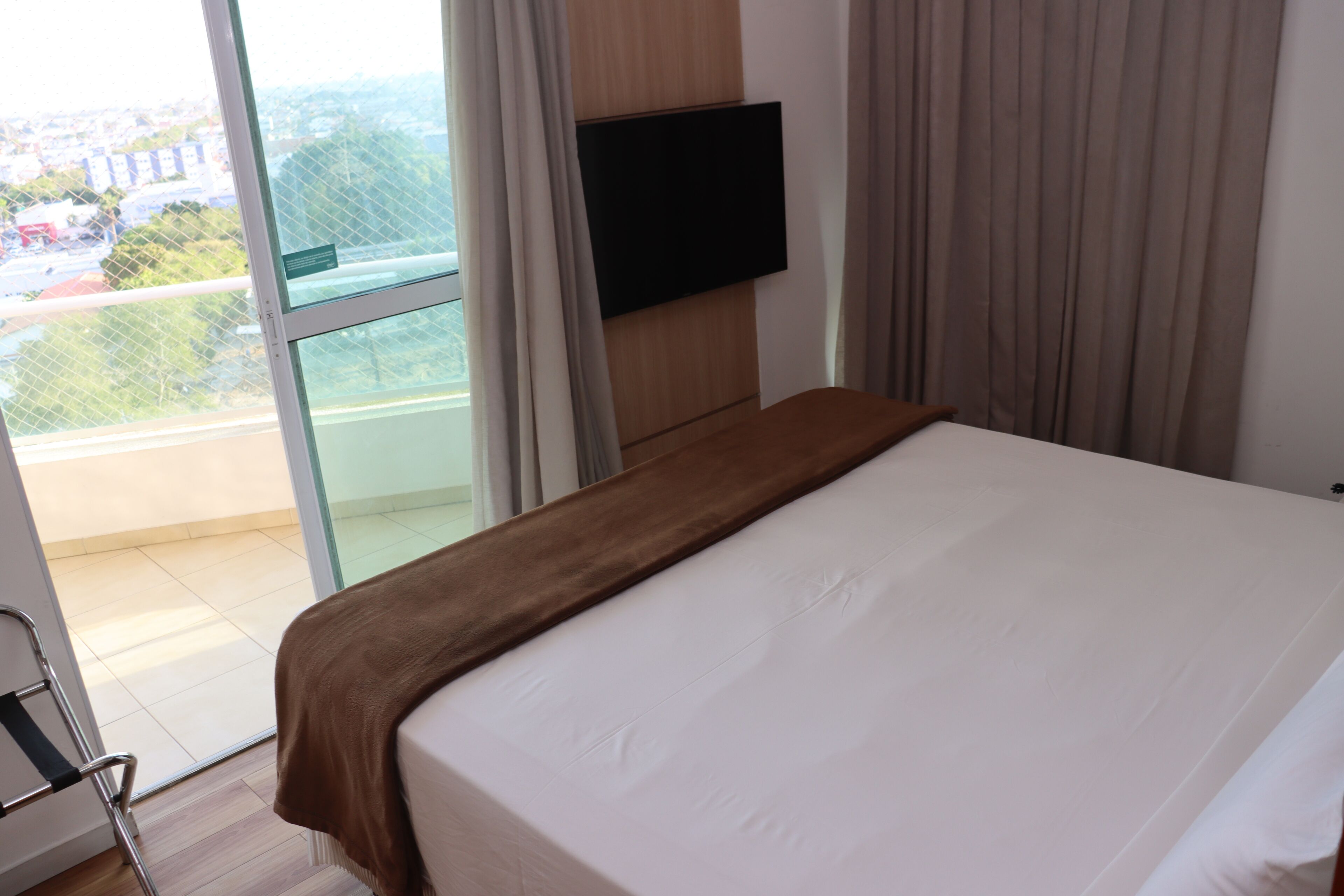 A hotel room with a bed, a TV mounted on the wall, and a view of the city through a large window.
