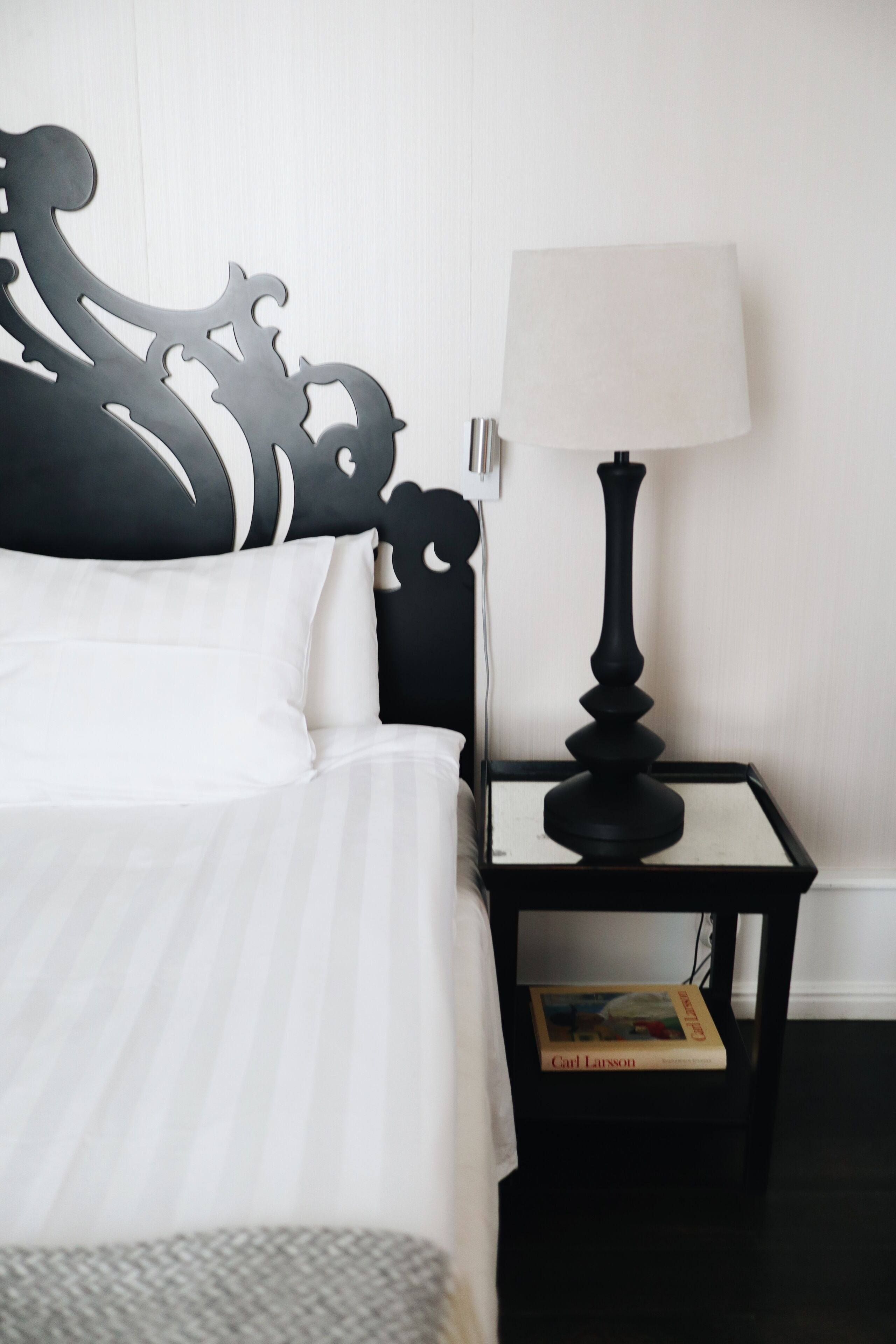A neatly made bed with white bedding and a black headboard, a black nightstand with a lamp and a book, and a plain white wall.
