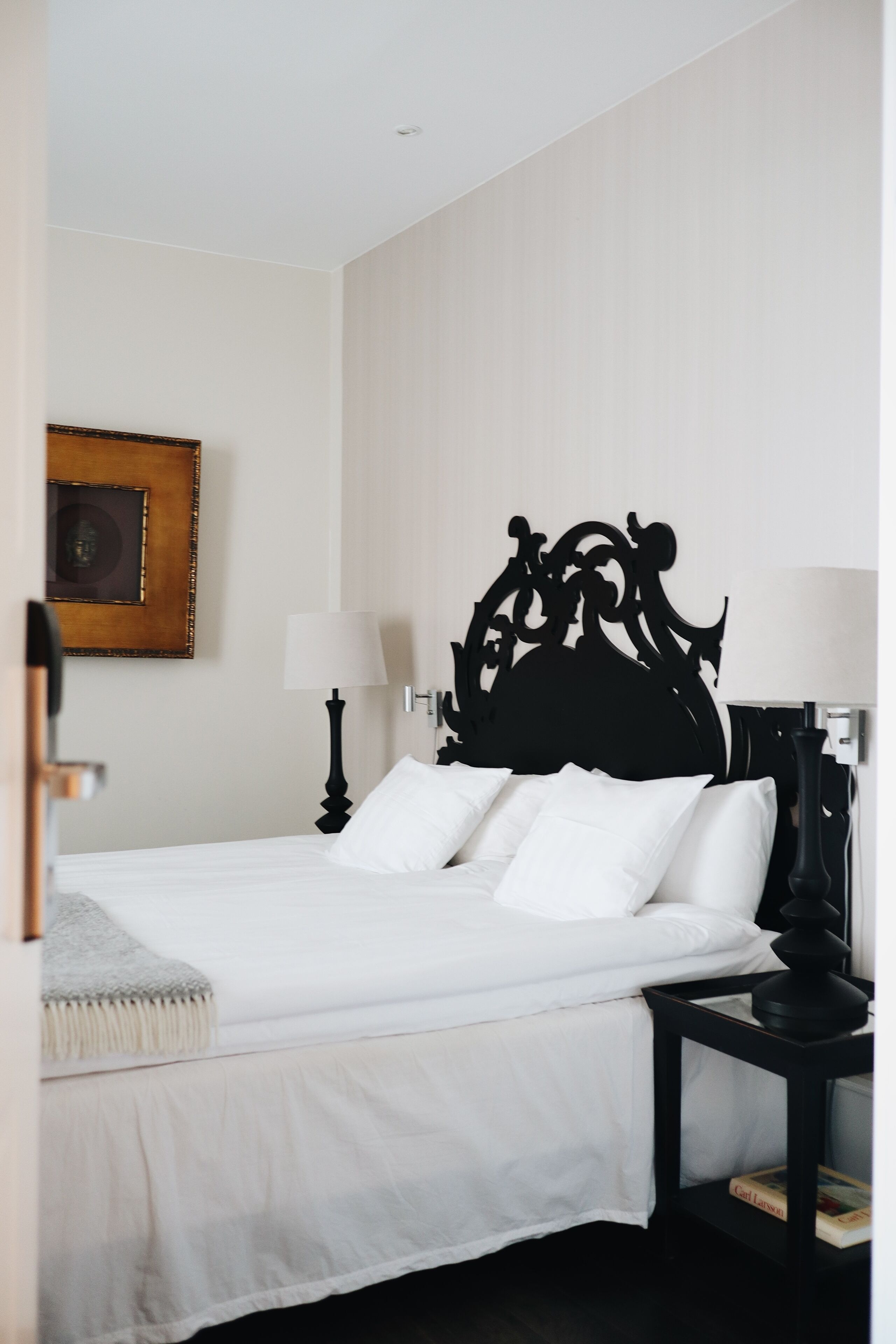 A neatly made bed with white linens and a black headboard, a bedside lamp, and a framed picture on the wall.